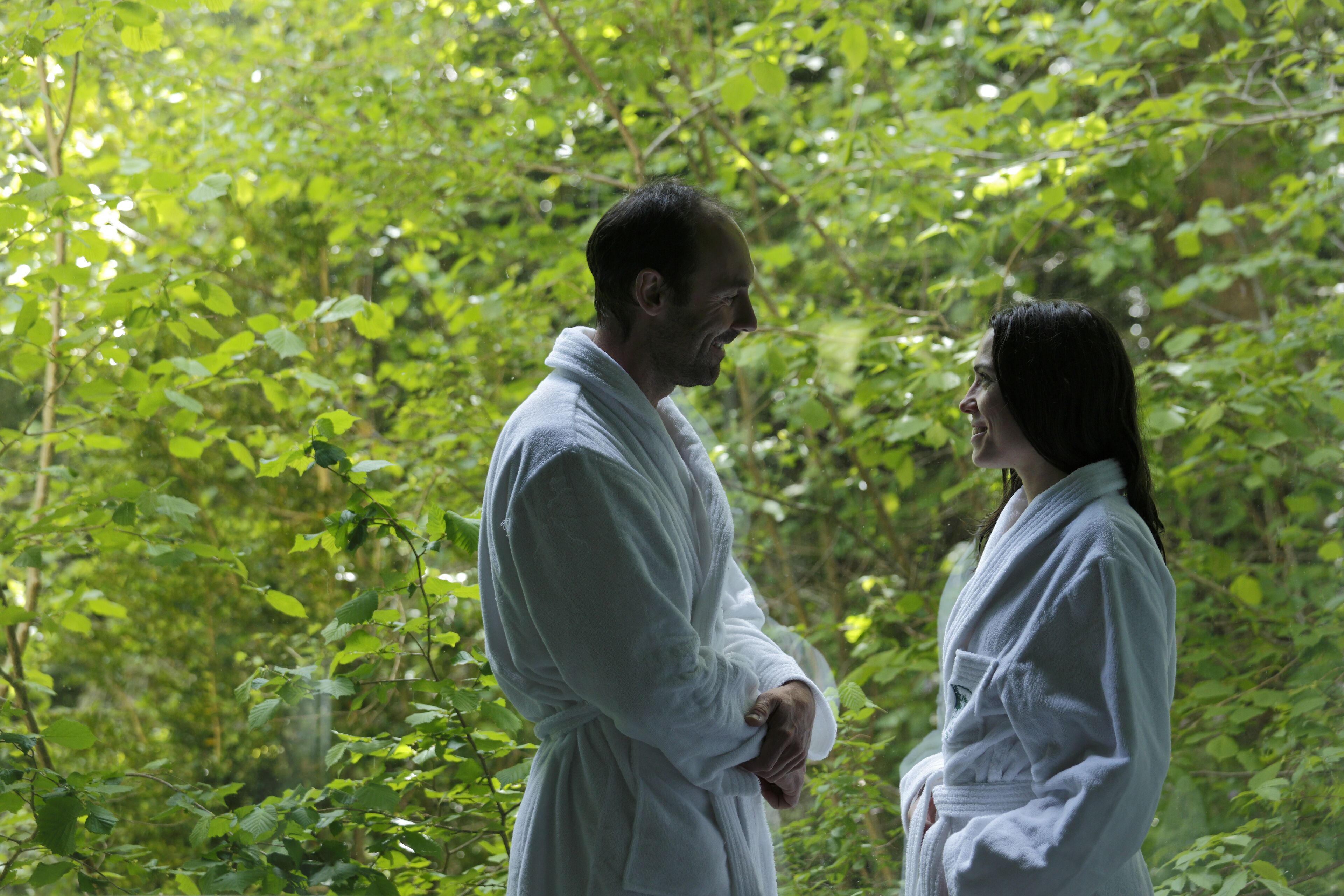 Two individuals in white robes standing in a lush, green forested area.