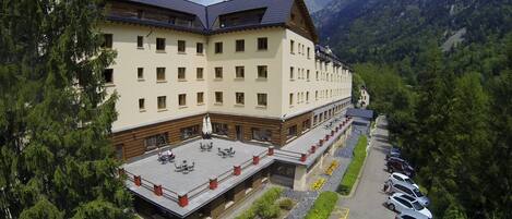 Un hotel de varios pisos con terraza en la azotea y aparcamiento, con montañas al fondo.