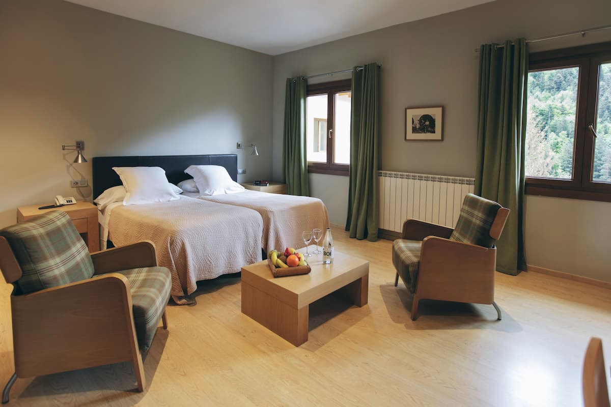 Quarto de hotel com duas camas, um espaço de estar com cadeiras, uma mesa pequena com frutas e copos, e uma janela com cortinas.