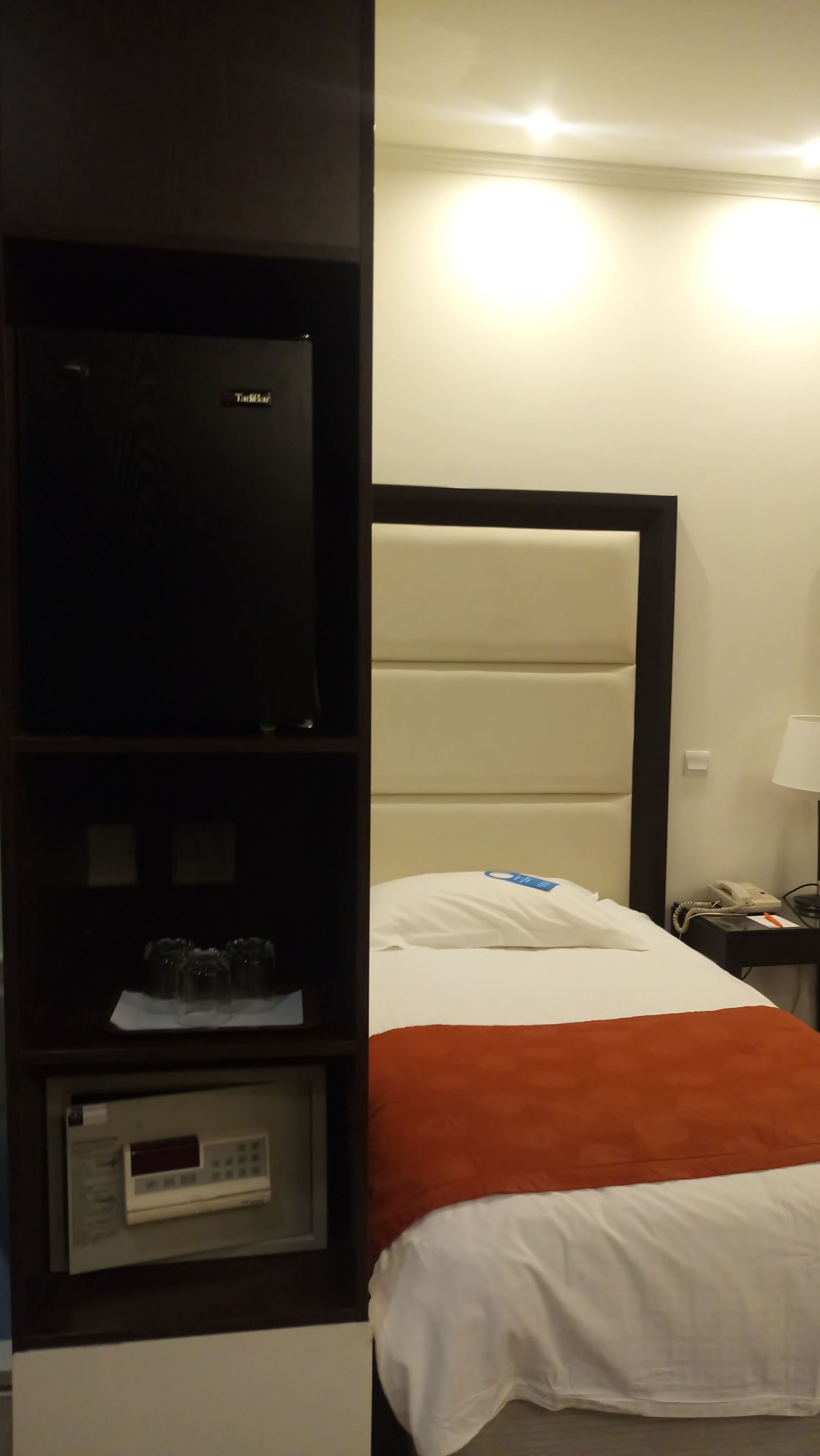 A hotel room with a bed, a TV on a shelf, a telephone, and a lamp.