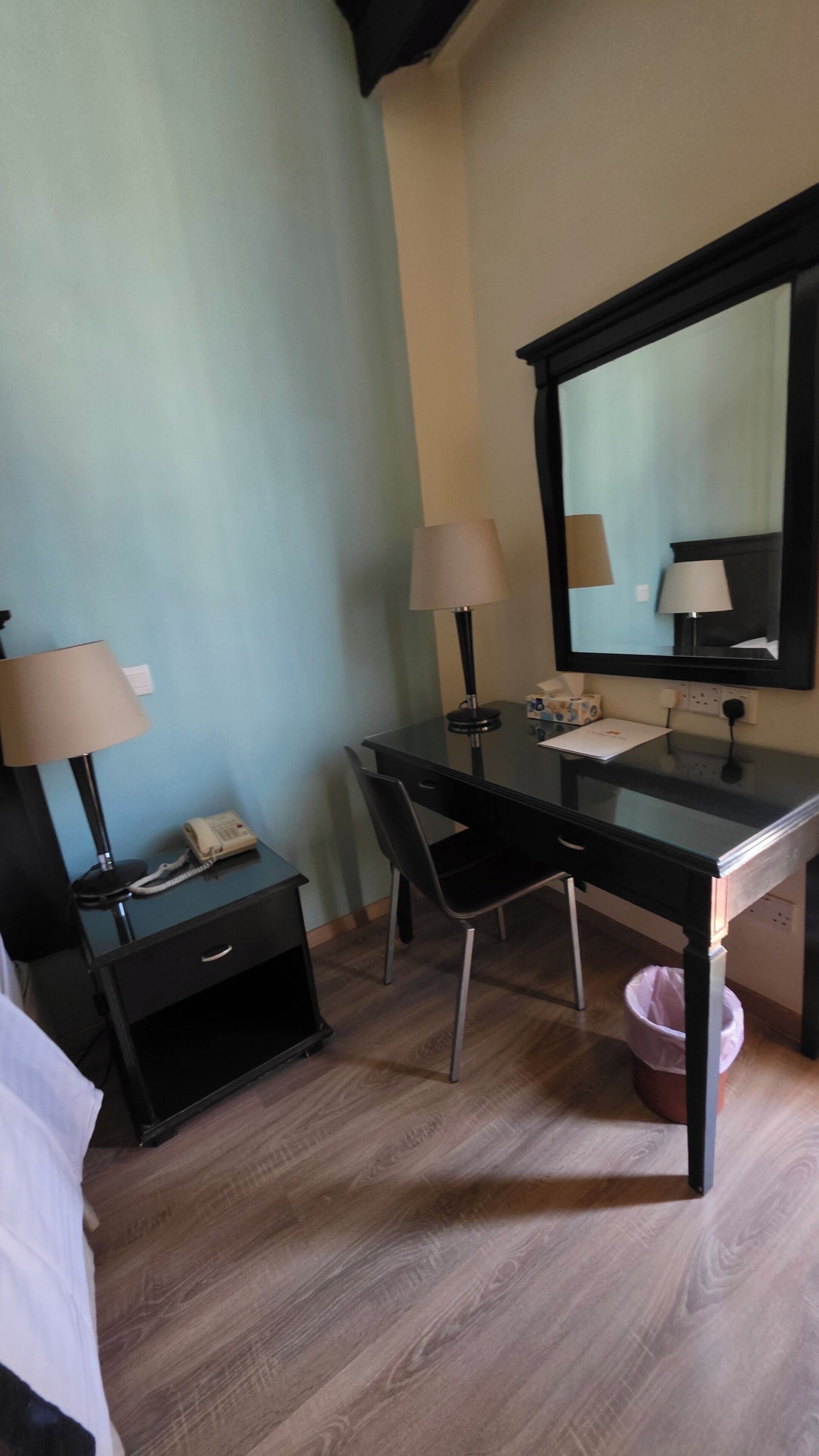 A room with a desk, two lamps, a telephone, a mirror, a chair, a small trash bin, and a bed.