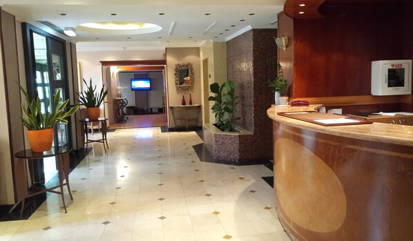 A hotel lobby with a reception desk, potted plants, a television, and a tiled floor. - Castelli Hotel (Nicosia)