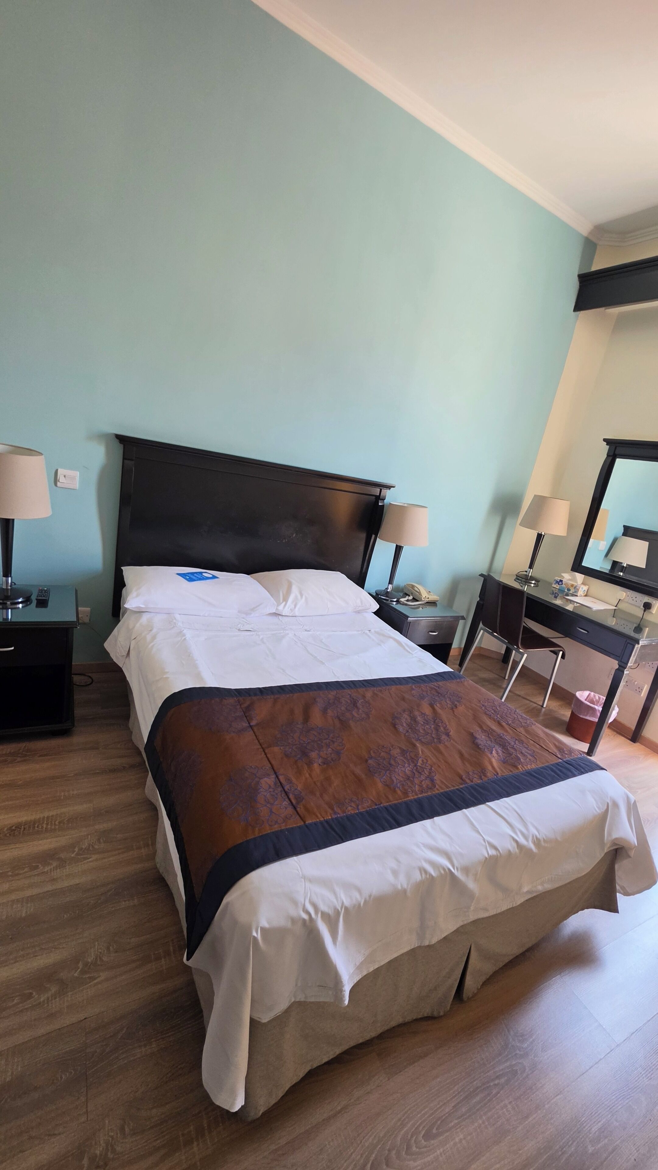 A neatly made bed with a dark headboard, a bedside table with a lamp, a desk with a chair, and a mirror.