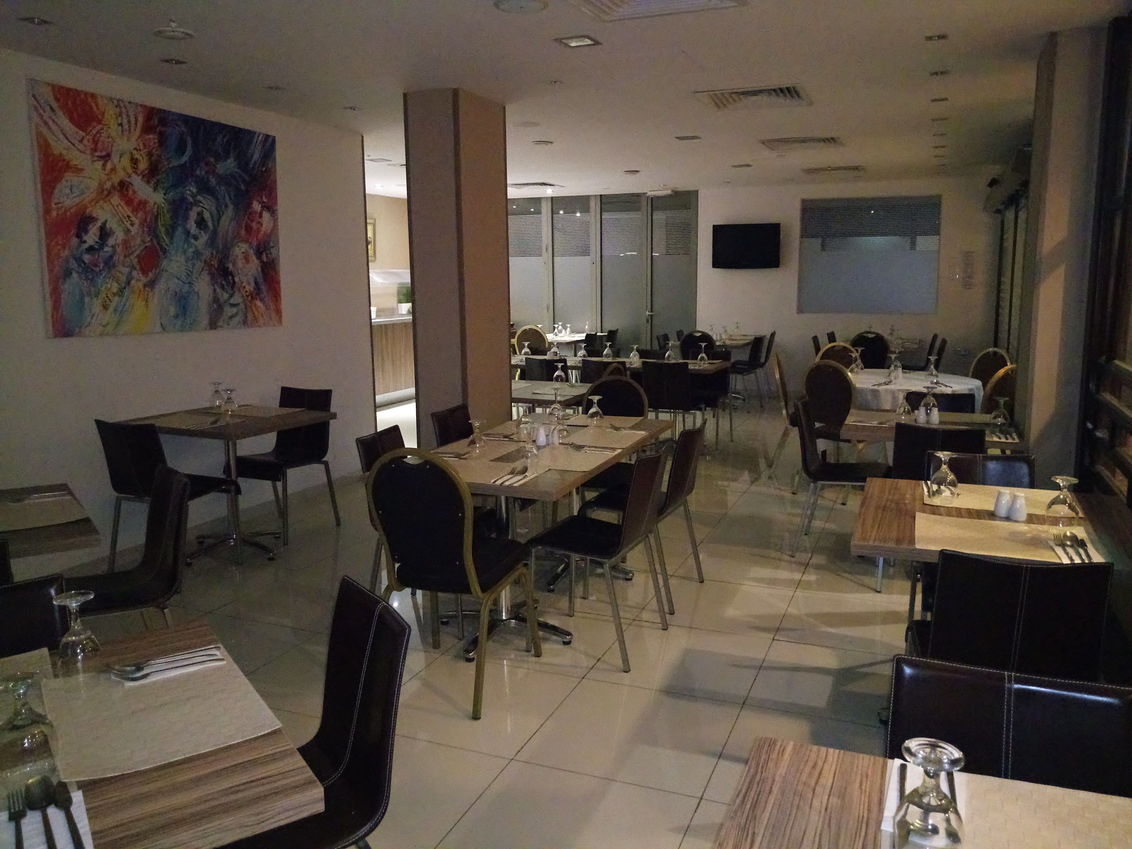 A modern dining area with multiple tables and chairs, a large abstract painting on the wall, and a television mounted on the wall.