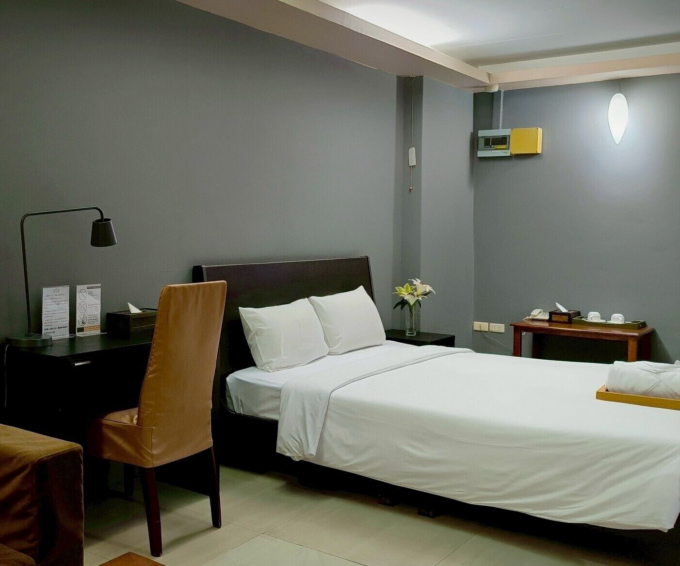 A hotel room with a bed, a desk with a lamp, a chair, a bedside table with a phone, and a small table with a vase of flowers.