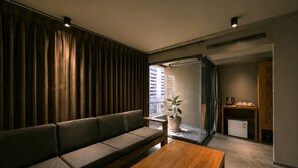 A modern living room with a sofa, a wooden coffee table, a potted plant, a kitchen area with a refrigerator, and a view of buildings outside. - Centric Place Hotel (Bangkok)