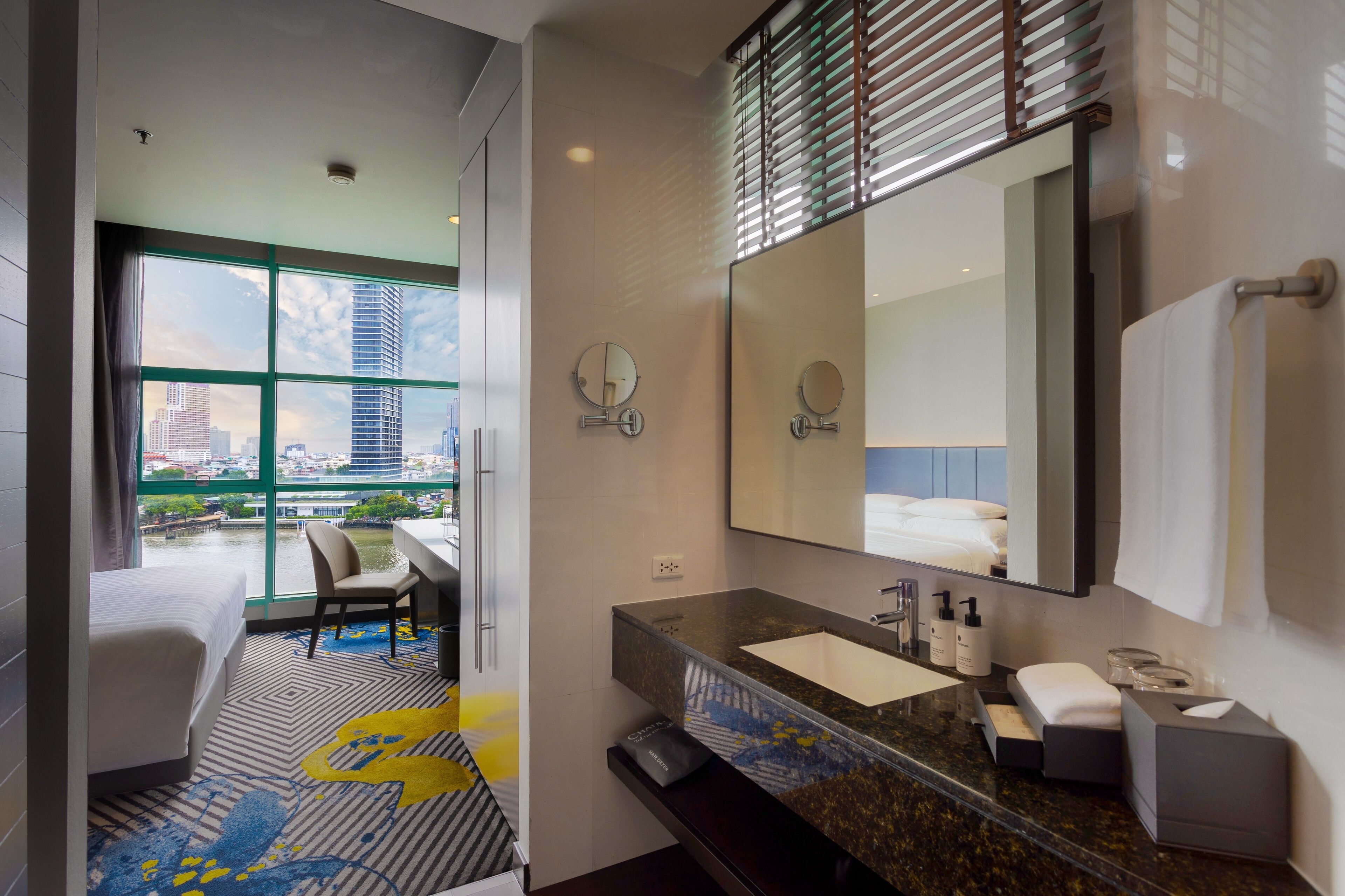 A modern hotel room with a large window, a bed, a desk, and a bathroom with a sink and mirror.