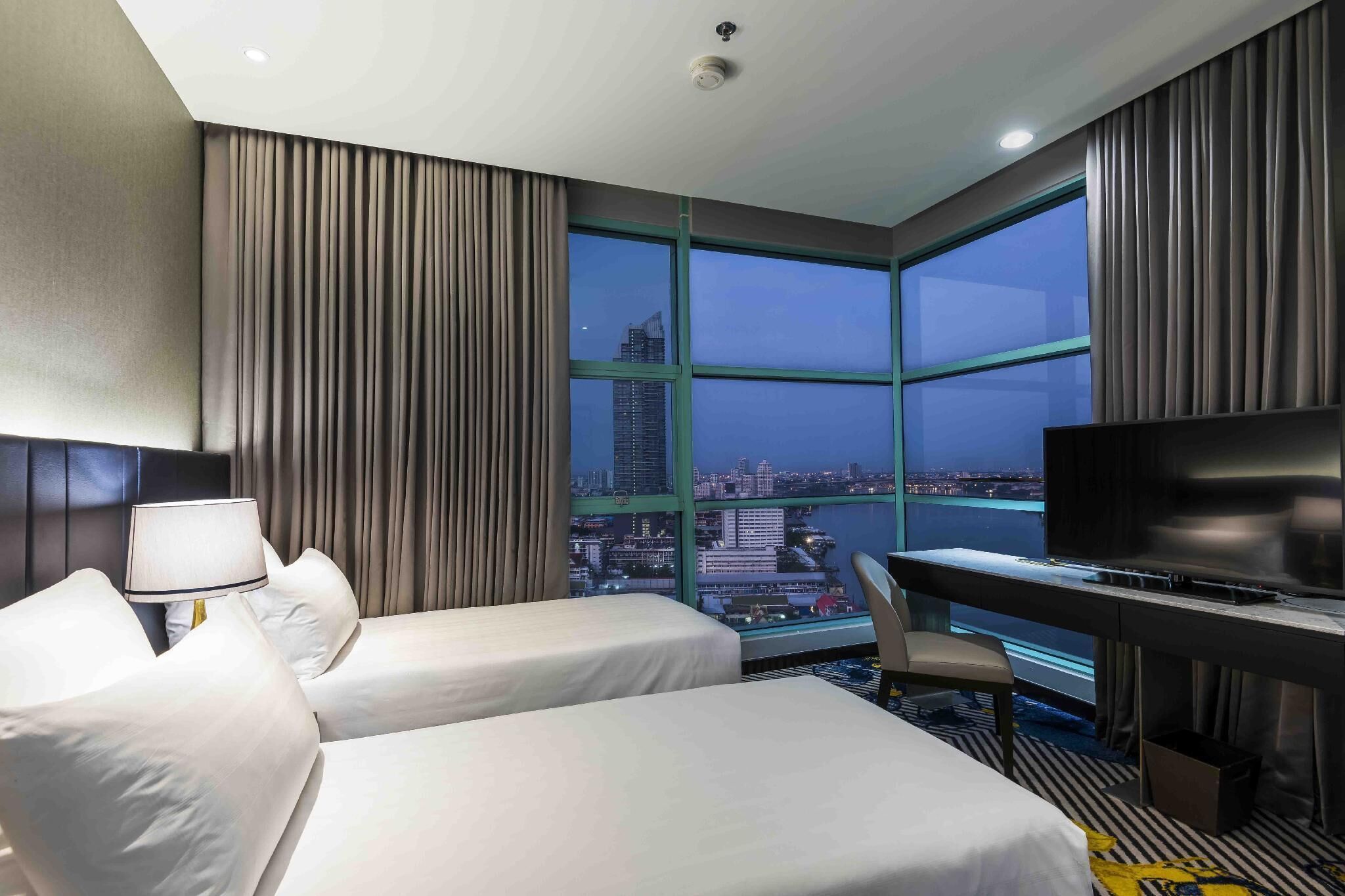 A hotel room with two beds, a desk, a television, and a large window offering a city view.