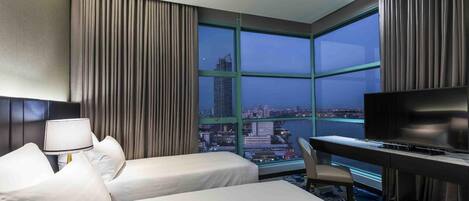 A hotel room with two beds, a desk, a television, and a large window offering a city view.