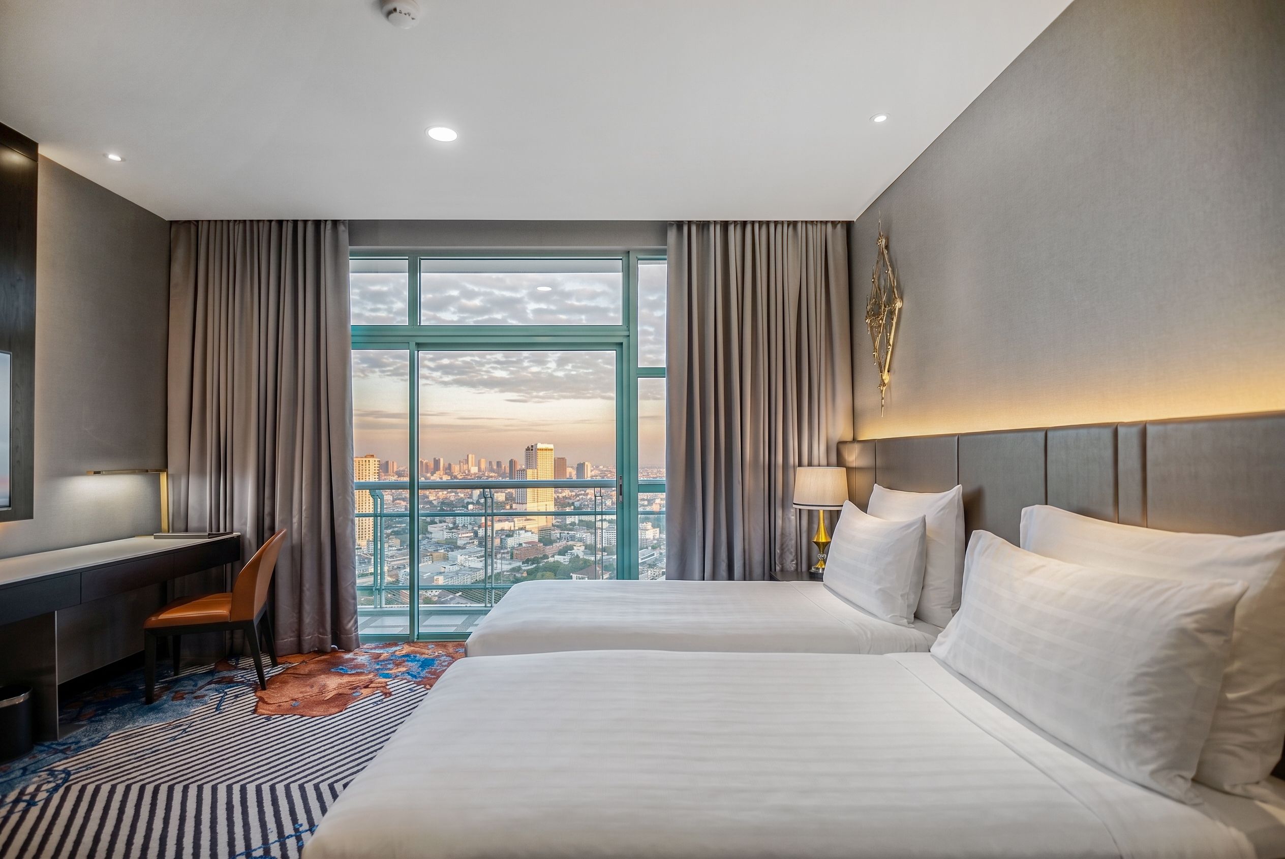 A hotel room with two beds, a desk, and a view of a cityscape.