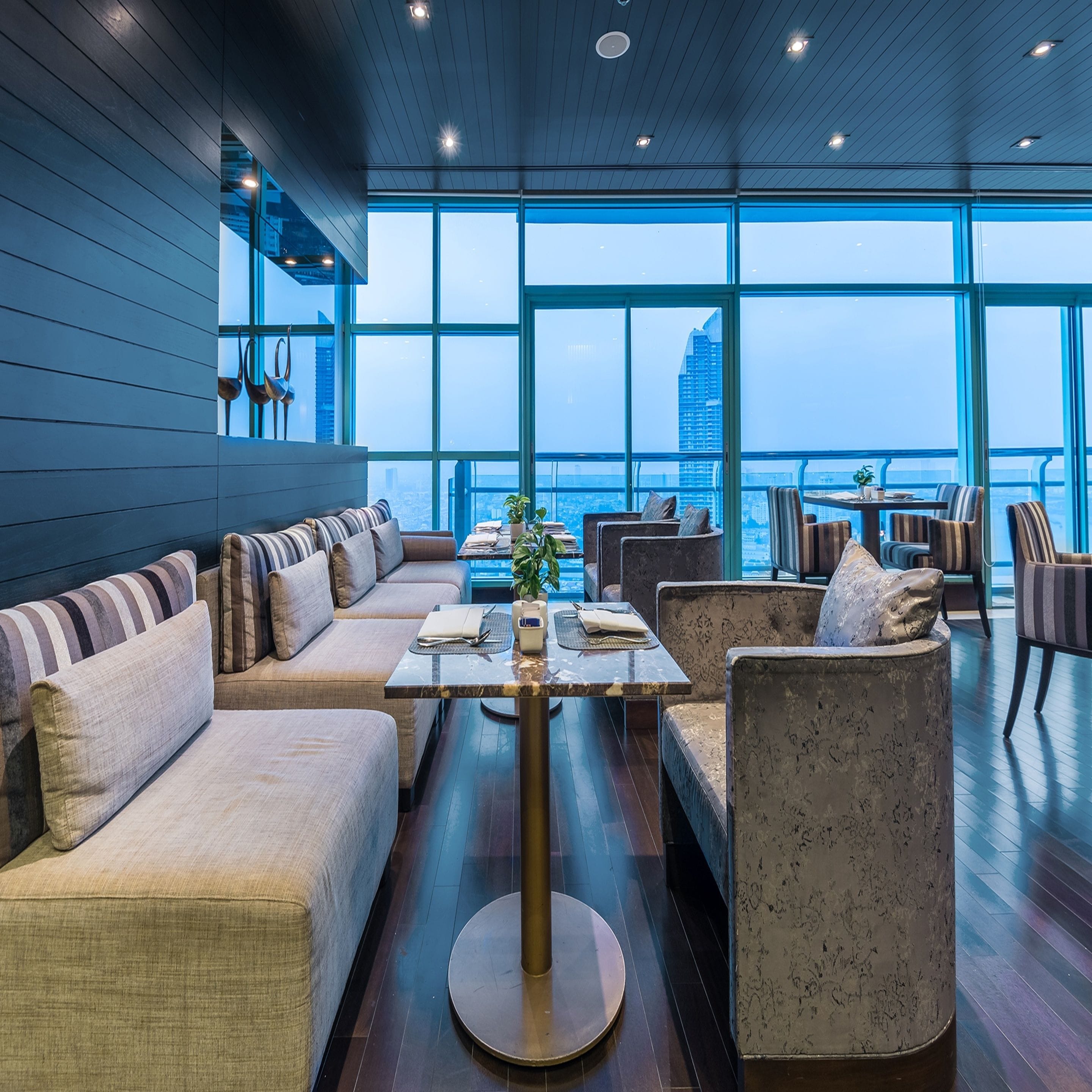 A modern lounge area with a sofa set, a coffee table, and chairs. Large windows offer a view of the cityscape.