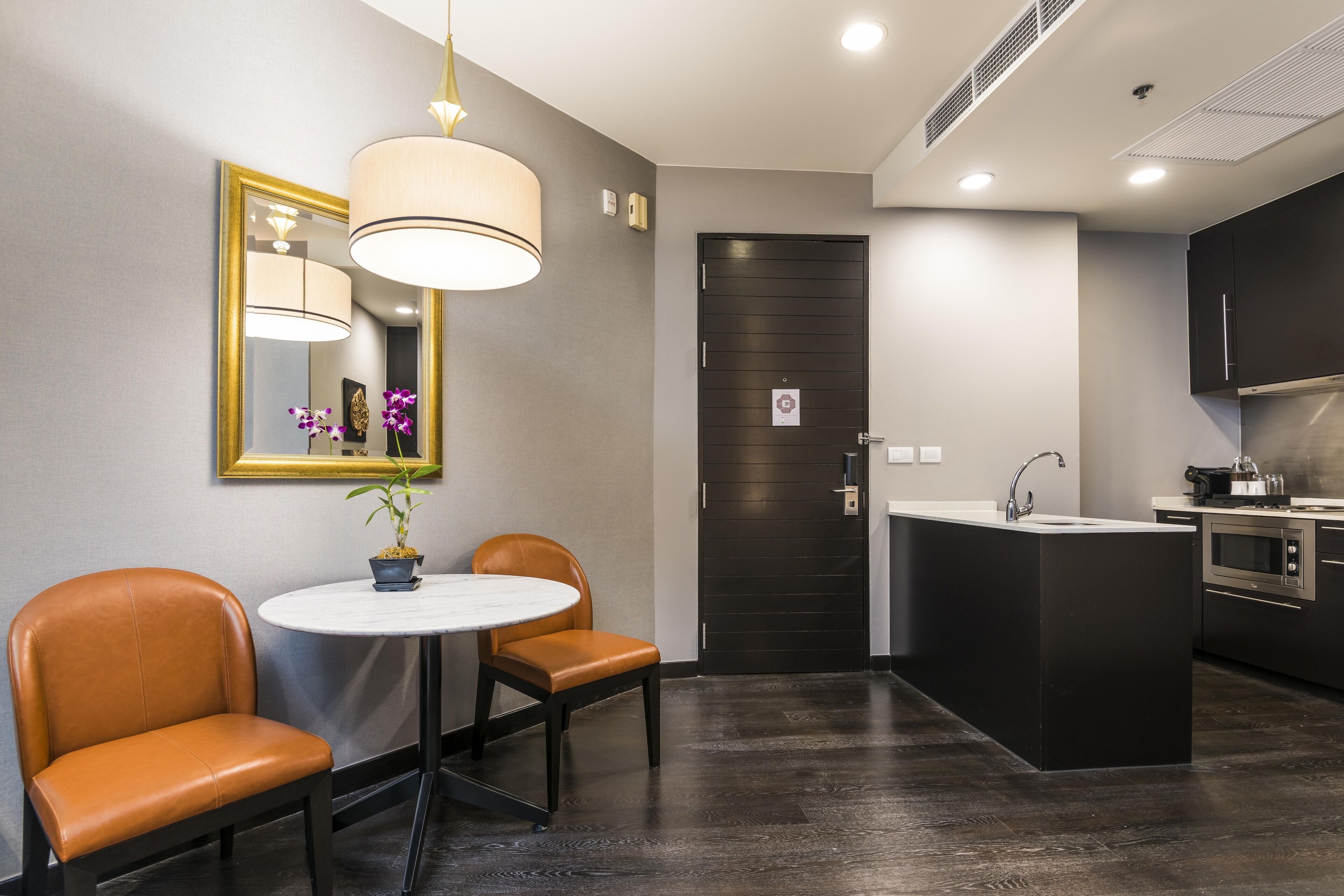 Una cocina moderna con suelo de madera oscura, una mesa redonda con dos sillas, una lámpara colgante y una puerta negra con un cartel pequeño.