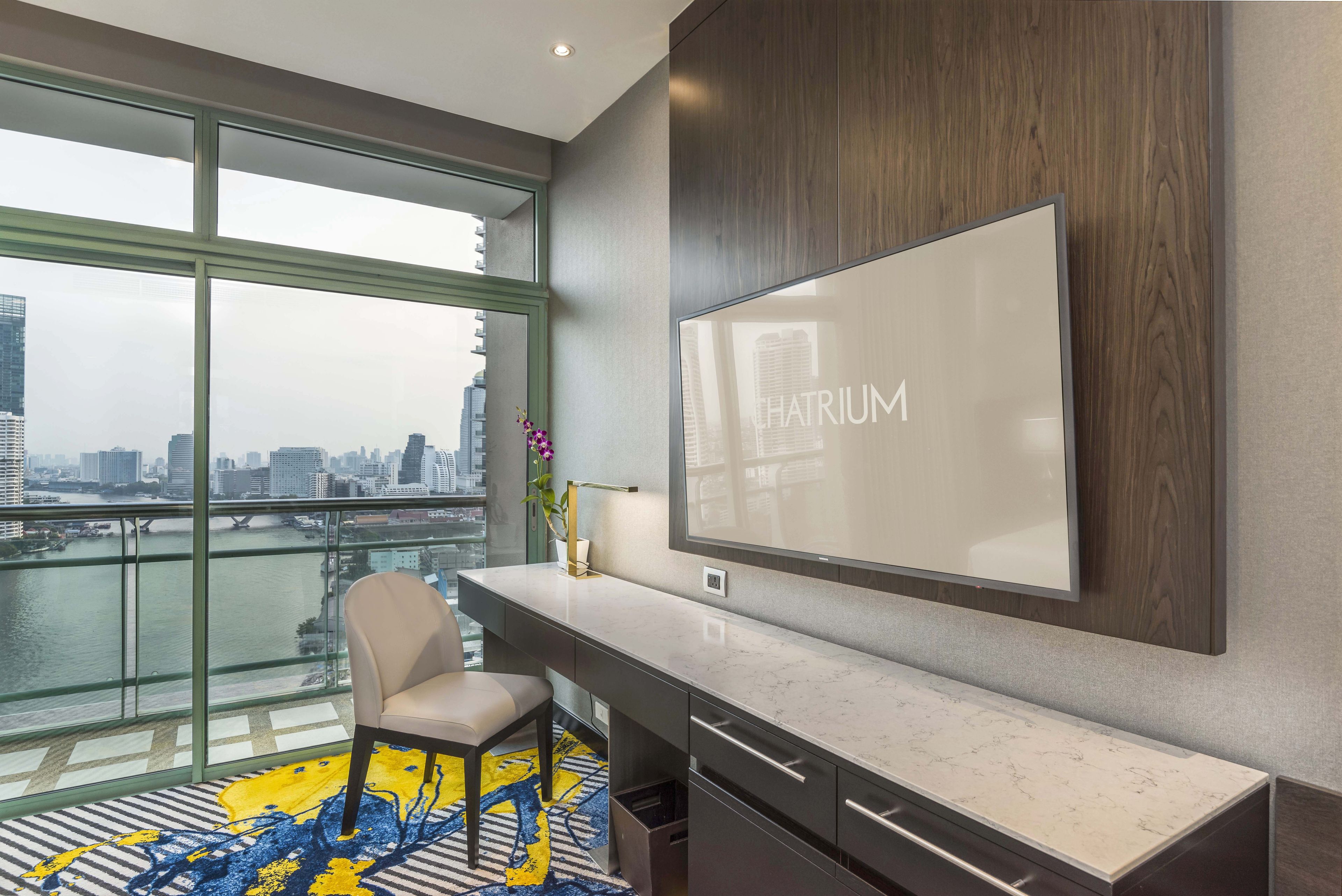 A modern hotel room with a large TV, a city view, a desk, and a chair.