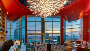 Vestíbulo de hotel de gran altura con un amplio ventanal que ofrece vistas a la ciudad, una lámpara de araña moderna y una zona de bar con piso estampado.