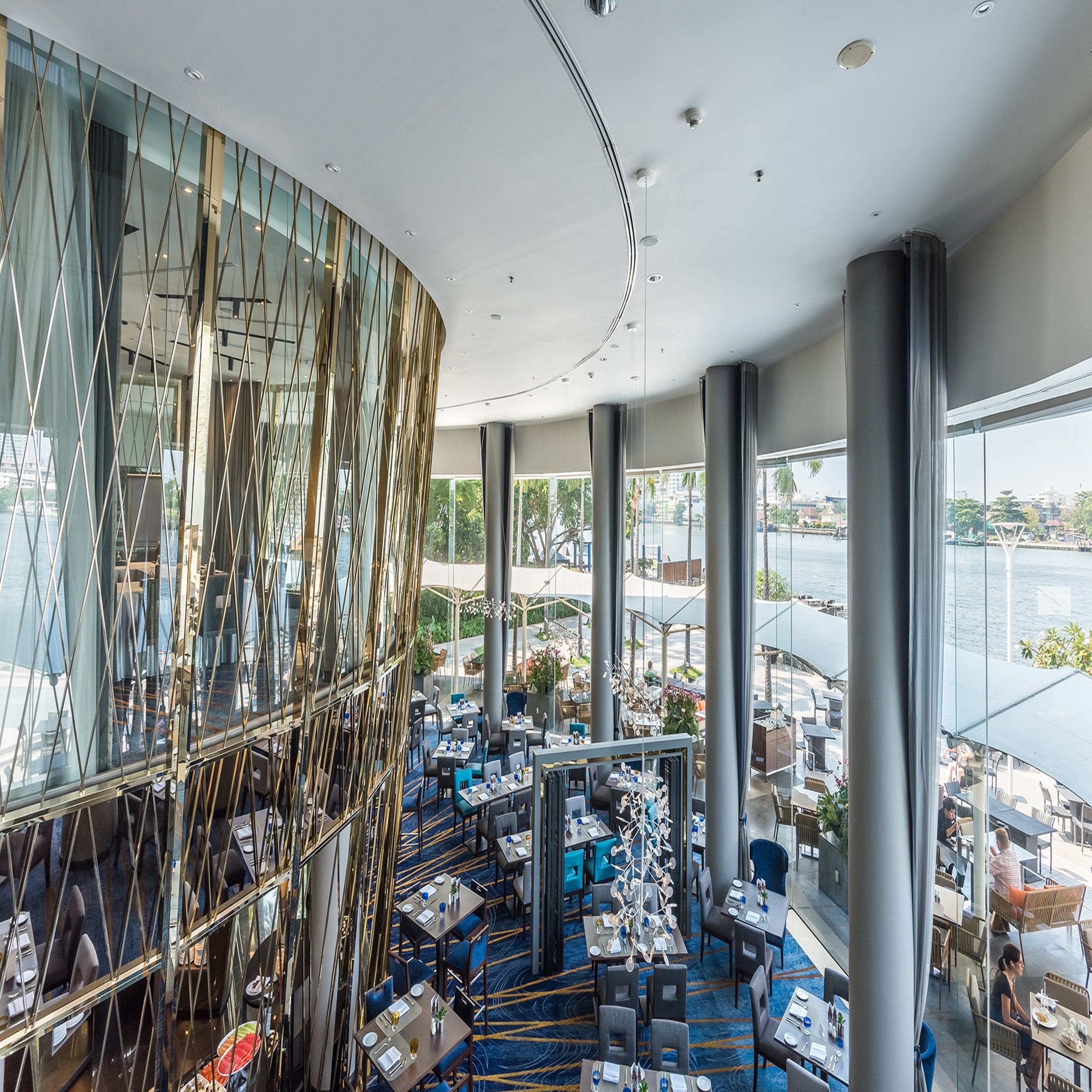 Un restaurante moderno con pared de vidrio curva, alfombra azul y mesas dispuestas para comer.