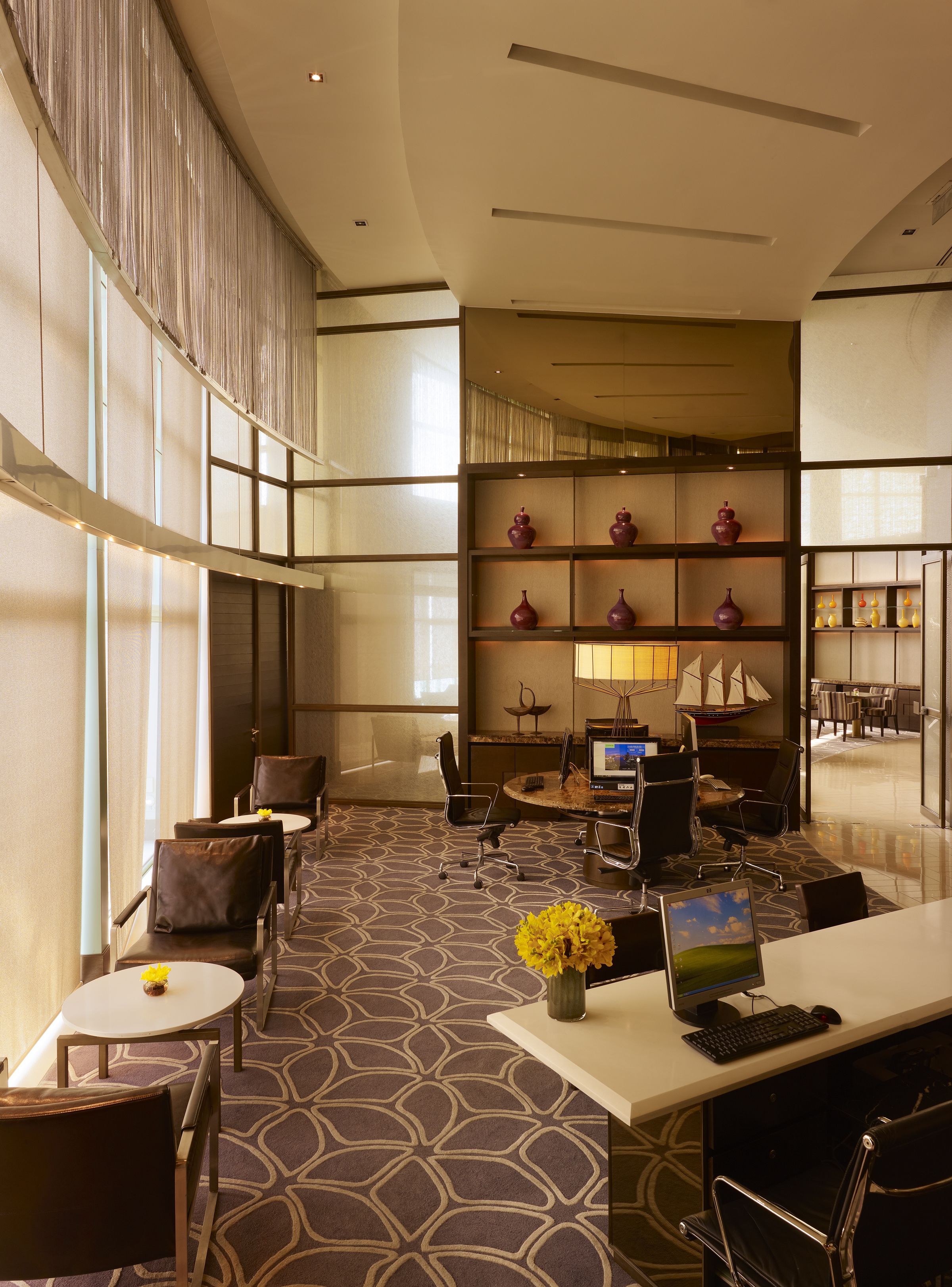 A modern hotel lobby with a high ceiling, large windows, a seating area with chairs and tables, a reception desk, and a decorative bookshelf.