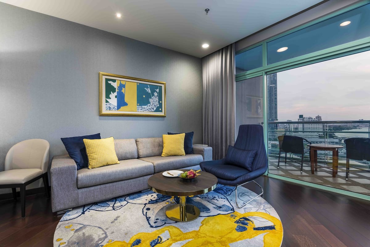 Une chambre d’hôtel moderne avec un canapé gris, un fauteuil bleu, une table basse ronde et une grande fenêtre donnant sur la ville.