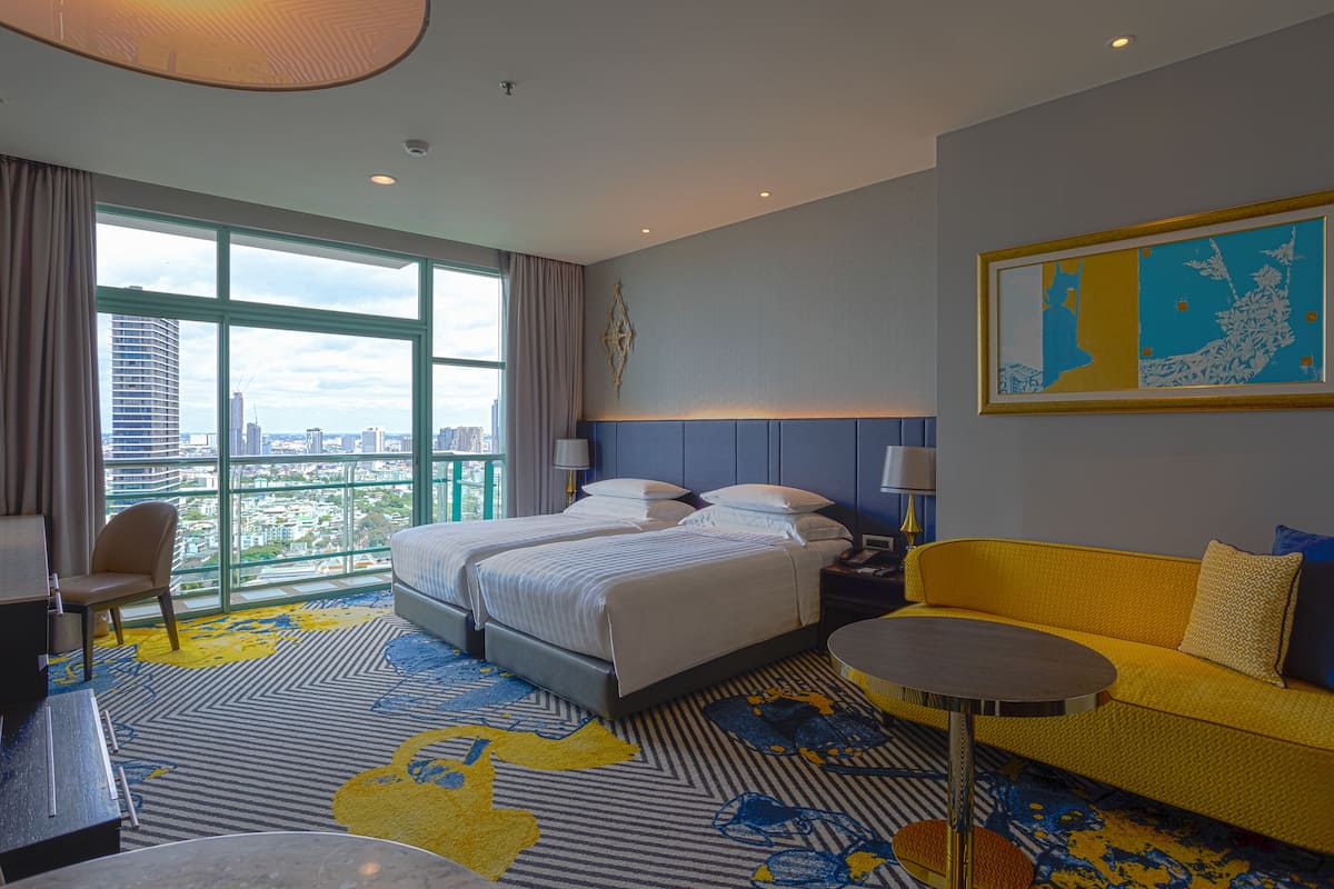 Une chambre d’hôtel avec deux lits, un canapé, un bureau et une grande fenêtre donnant sur la ville.