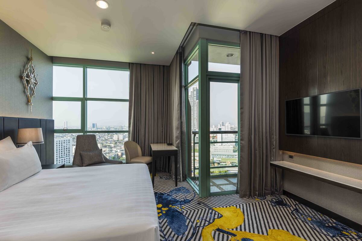 Une chambre d’hôtel avec un grand lit, un bureau, une chaise, une télévision et une vue sur la ville grâce à de grandes fenêtres.