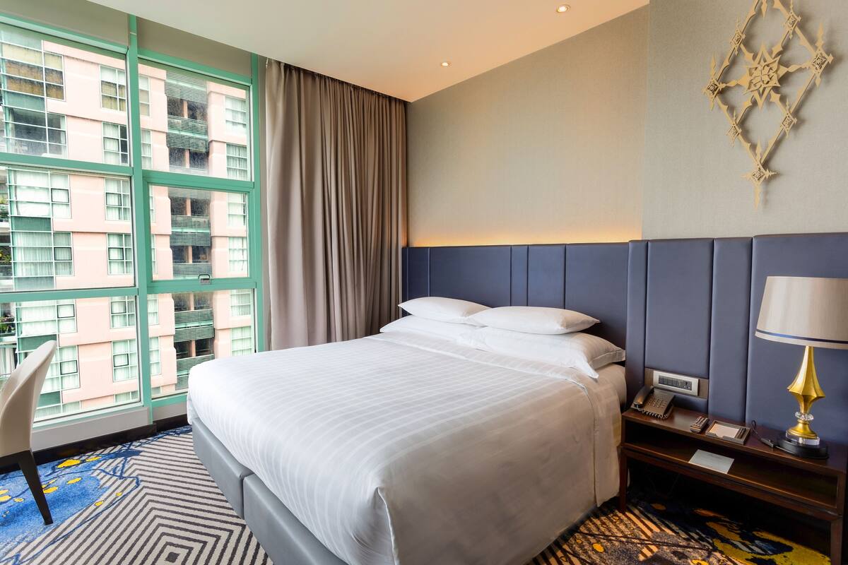 Une chambre d’hôtel avec un grand lit, un bureau avec une lampe, un téléphone et une vue sur un immeuble à travers de grandes fenêtres.