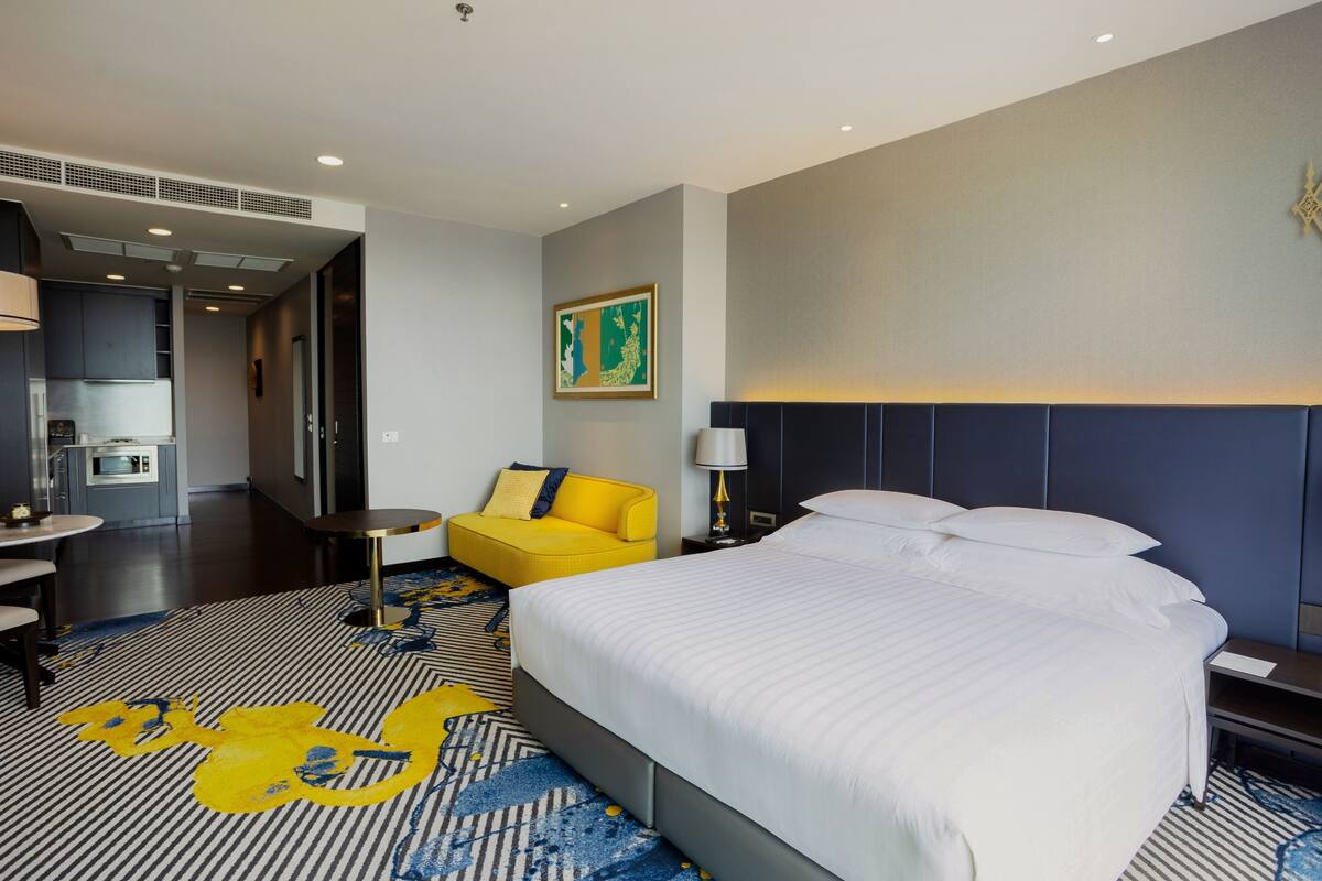 Une chambre d’hôtel avec un grand lit, un fauteuil jaune, une petite table et, au fond, une kitchenette.
