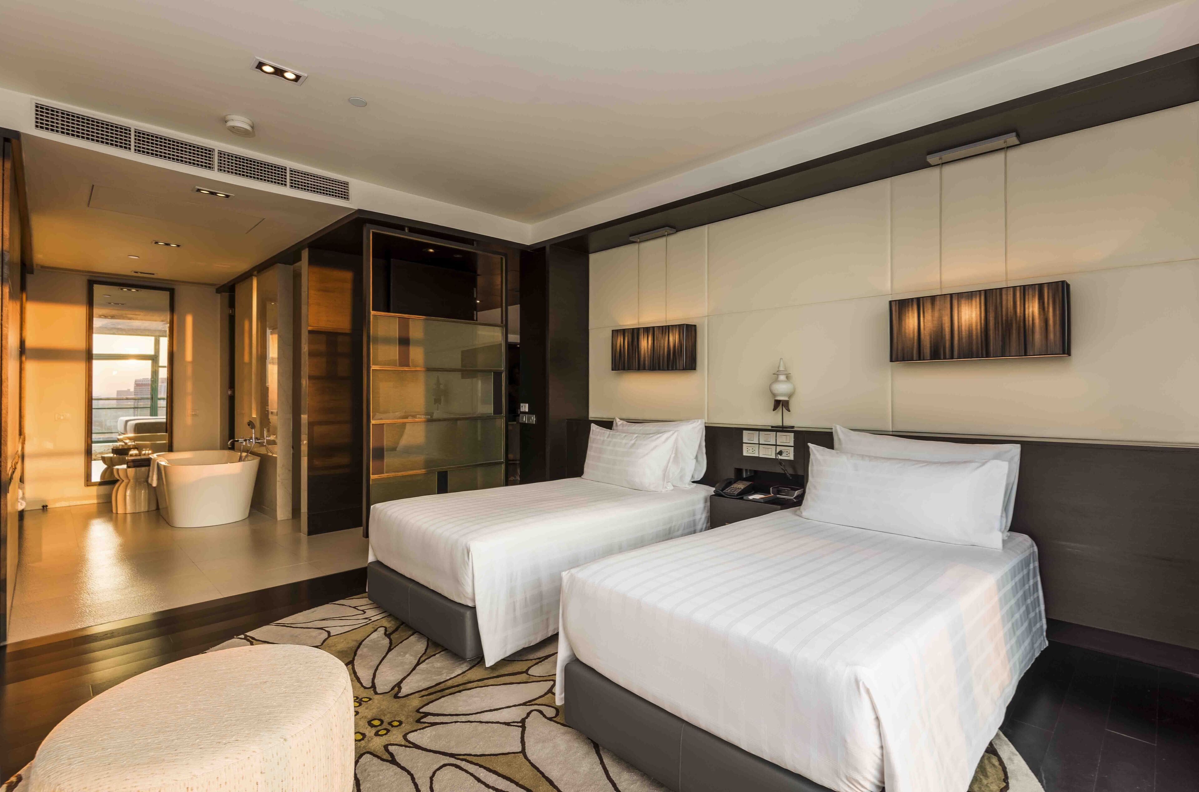 A modern hotel room with two beds, a bathtub, a large mirror, and a view of the city.