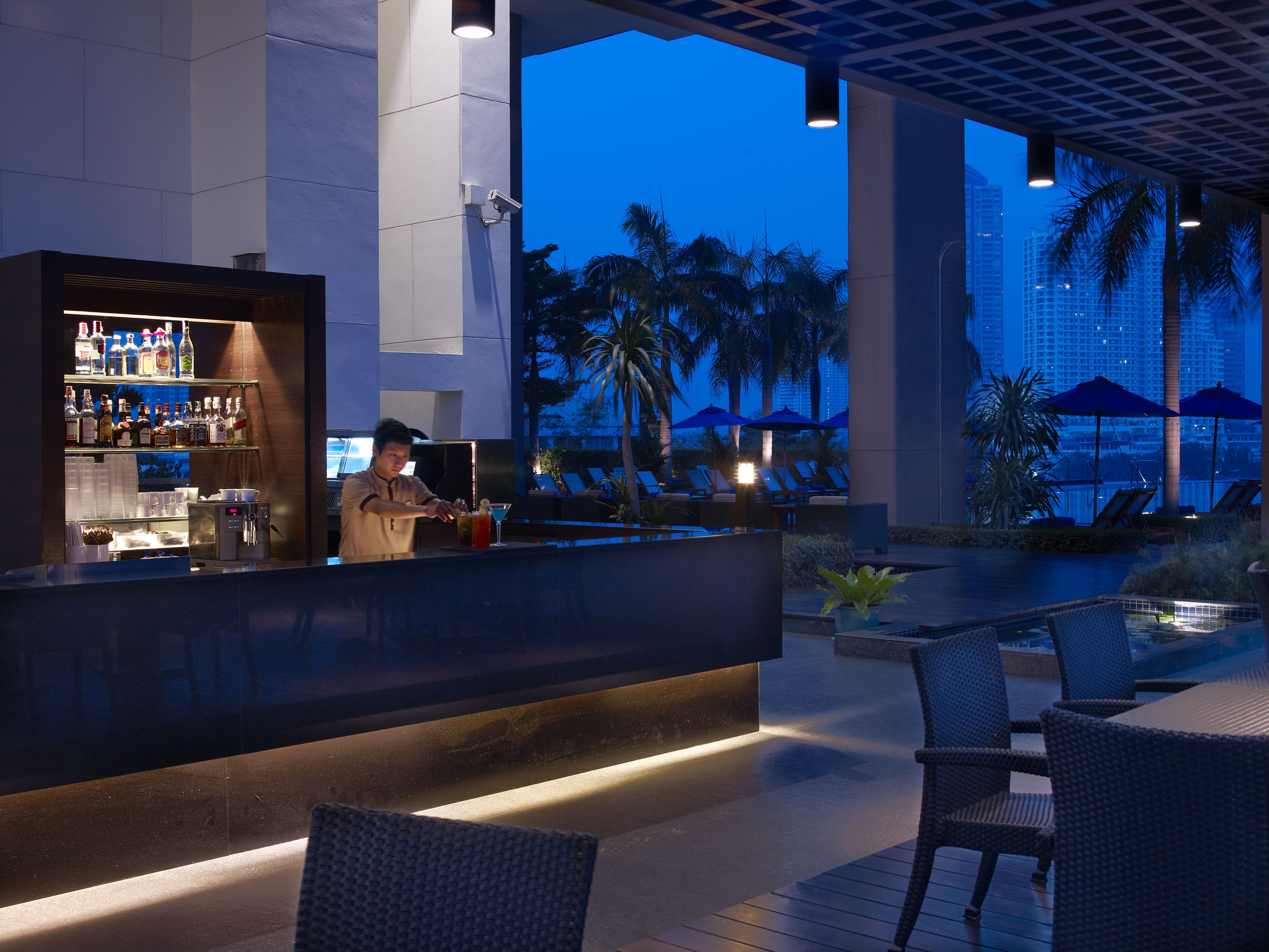 Una zona de bar con mostrador, estantes llenos de botellas y vistas a palmeras y edificios al fondo.