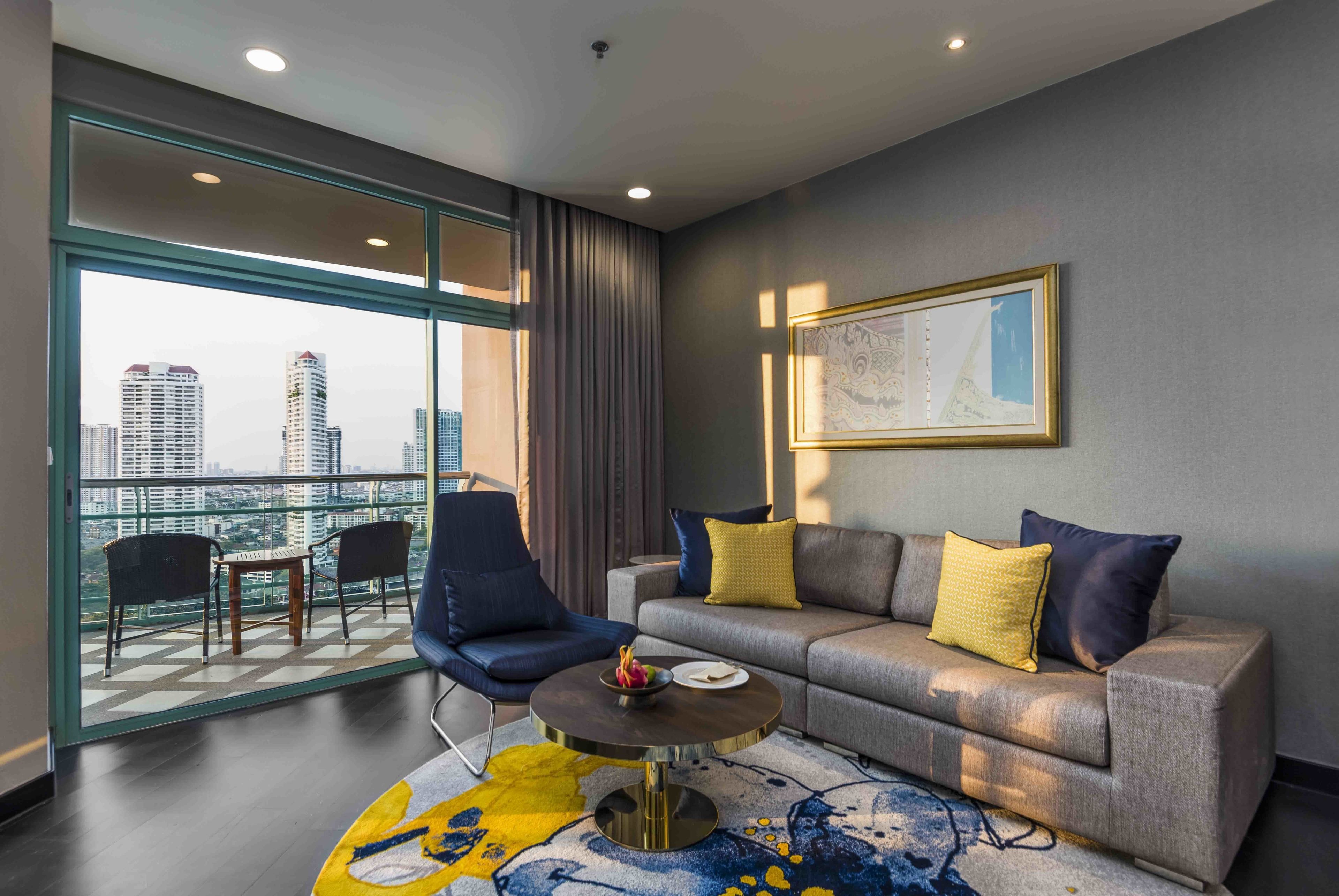 A modern hotel room with a balcony, a grey sofa with yellow and blue cushions, a round coffee table, and a city view.
