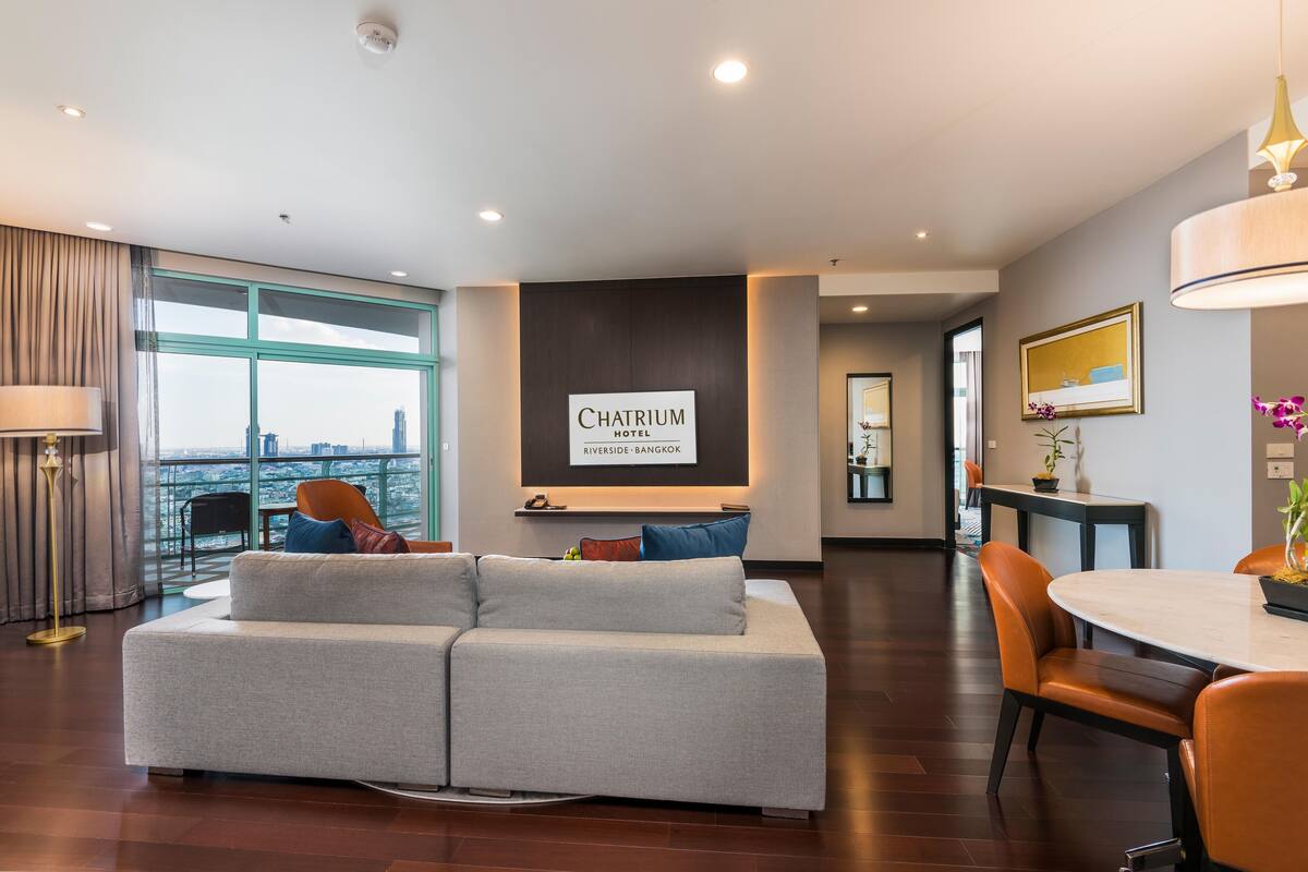 Une chambre d’hôtel moderne avec un canapé gris, une table à manger avec des chaises, une grande fenêtre donnant sur la ville et une enseigne indiquant « Chatrium Riverside – Bangkok ».
