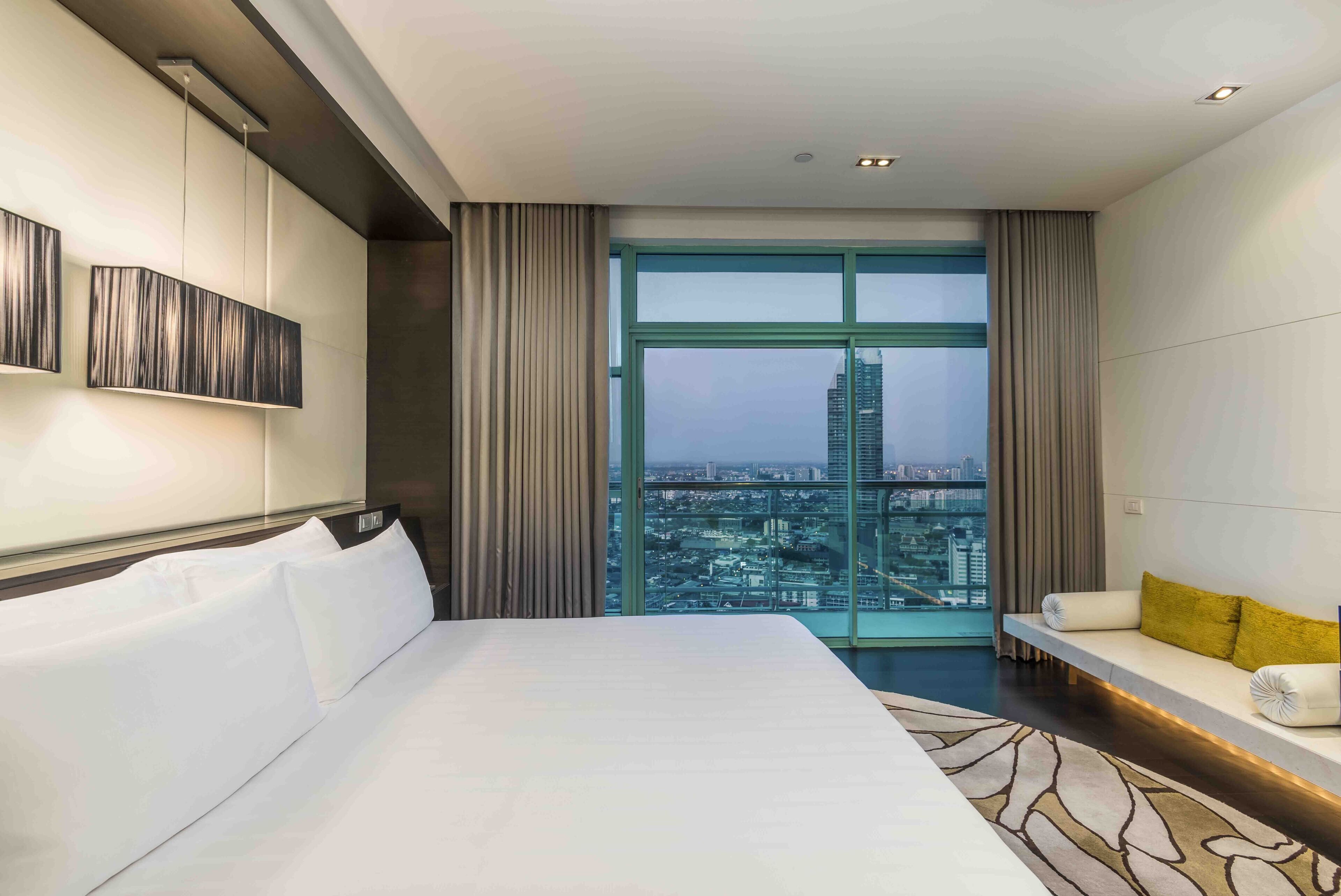 A modern hotel room with a large bed, a sofa, and a view of a cityscape through a large window.