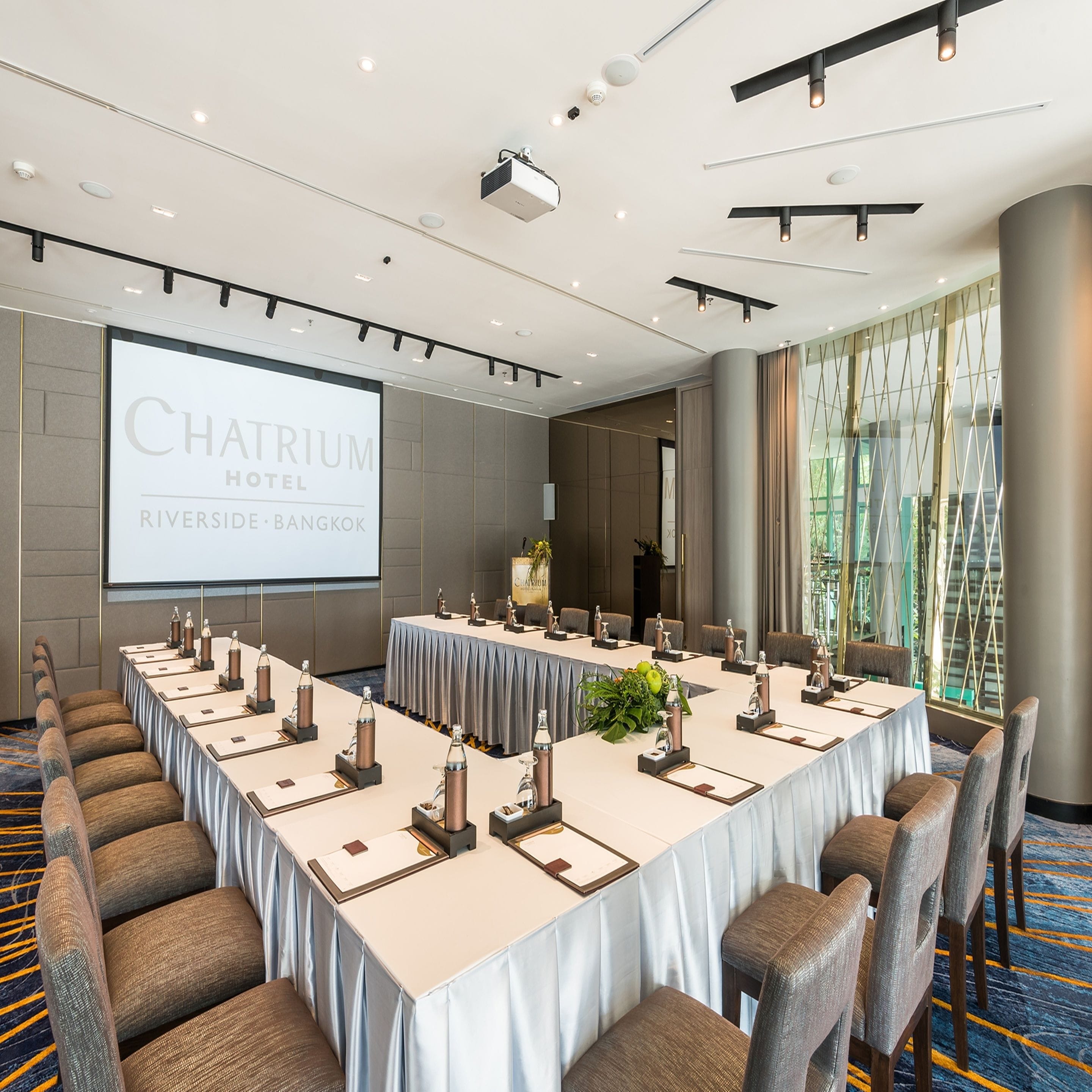 A conference room at Chatrium Hotel with a long table, chairs, a projector screen, and a large window.