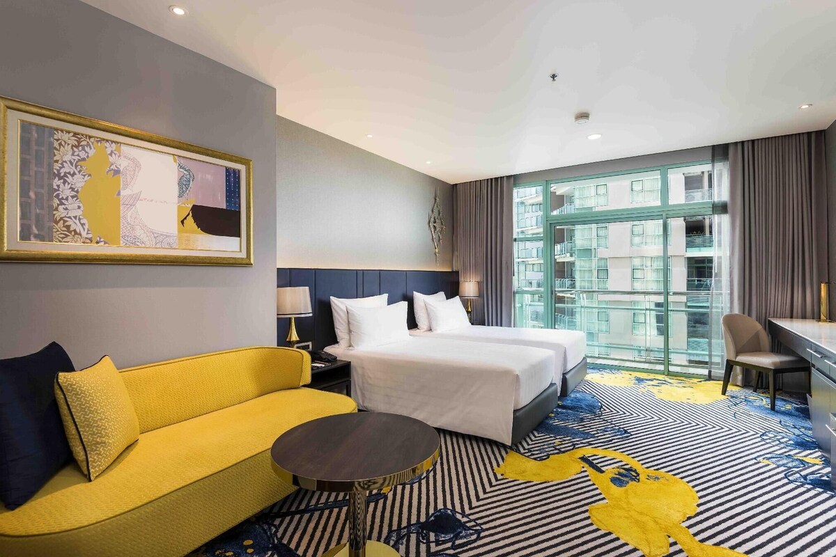 A hotel room with a bed, a yellow sofa, a small round table, and a large window with a view of buildings.