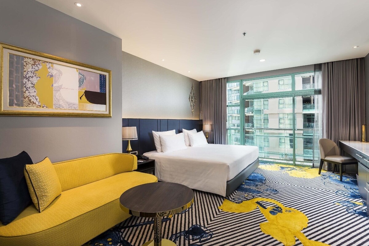 A hotel room with a bed, a yellow sofa, a round table, a desk, and a large window with a view of buildings.