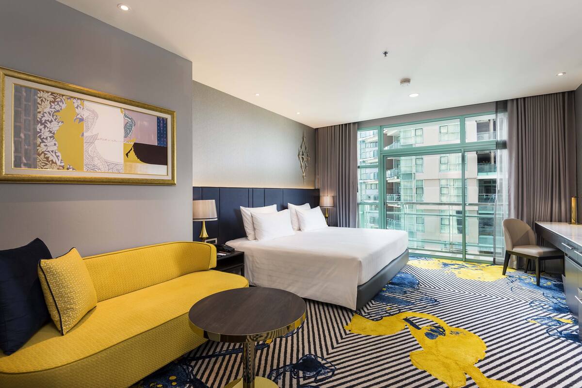 Une chambre d’hôtel avec un lit, un canapé jaune, un bureau et une grande fenêtre donnant sur des immeubles.