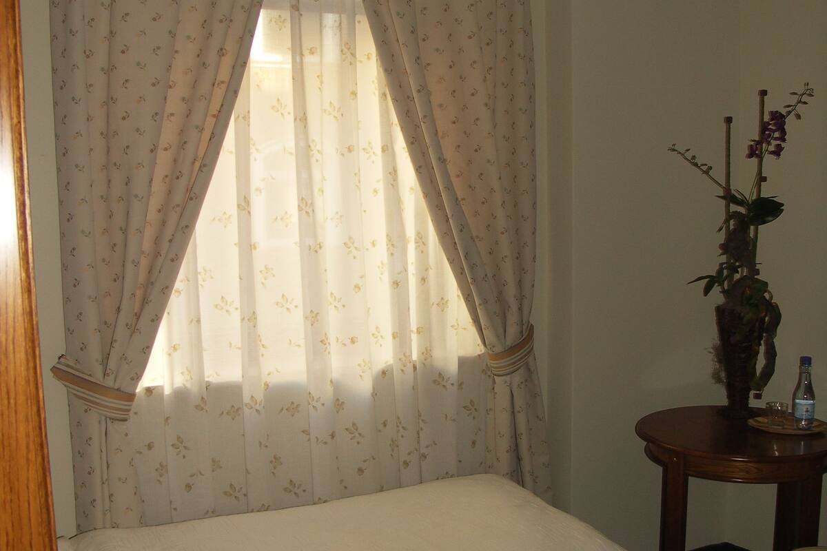 Ein Schlafzimmer mit einem Bett, einem Fenster mit durchscheinenden Vorhängen, einer Vase mit Blumen und einem kleinen Tisch mit einer Flasche.