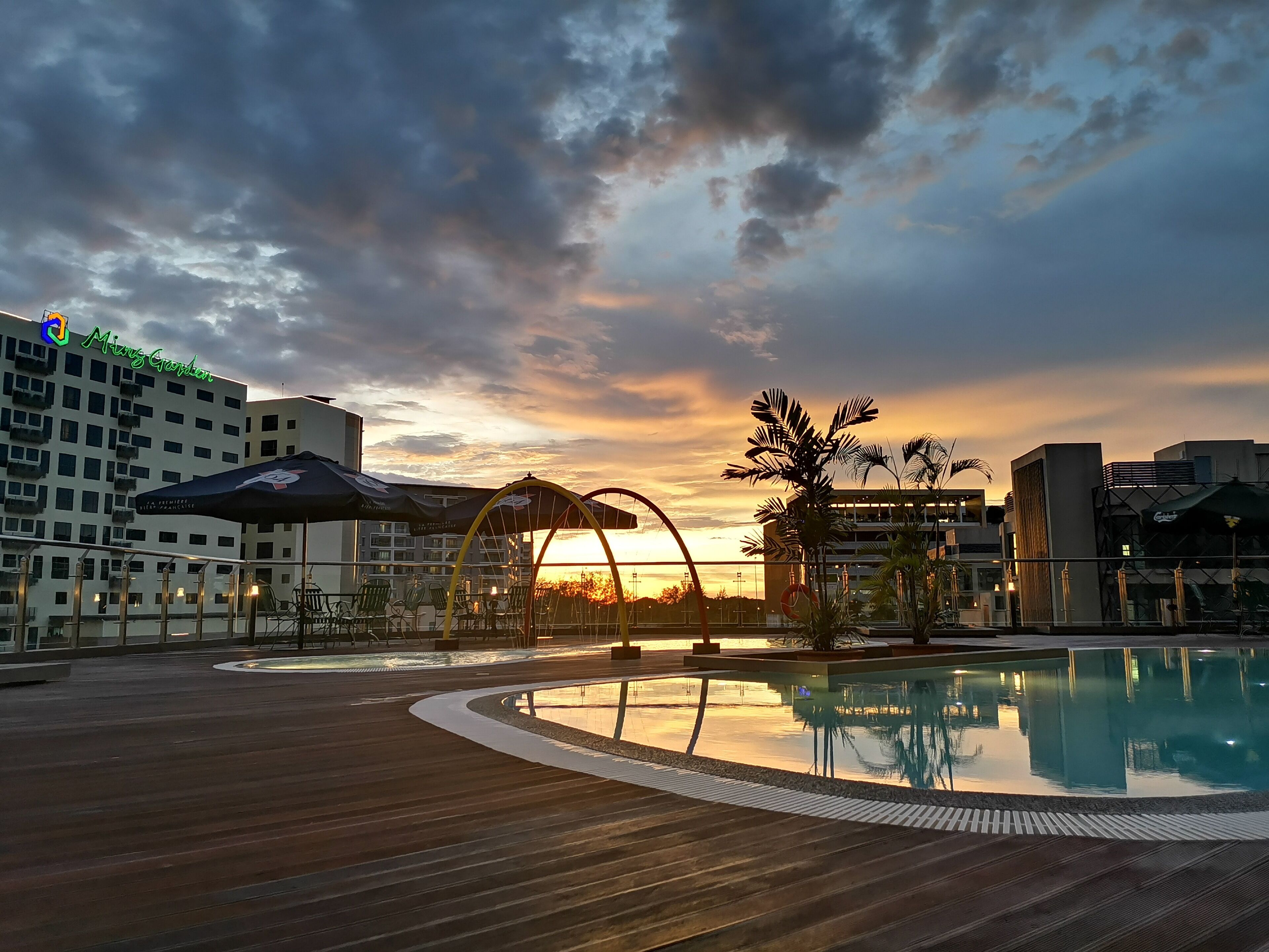 A rooftop pool area with a sunset view, outdoor seating, and a building with a green sign.