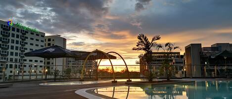 A rooftop pool area with a sunset view, outdoor seating, and a building with a green sign.