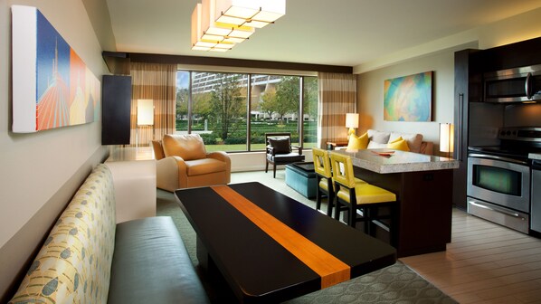 A modern hotel room with a sofa, dining table, chairs, a kitchen area, and large windows offering a view of the outdoors. - Bay Lake Tower at Disney's Contemporary Resort (Lake Buena Vista)
