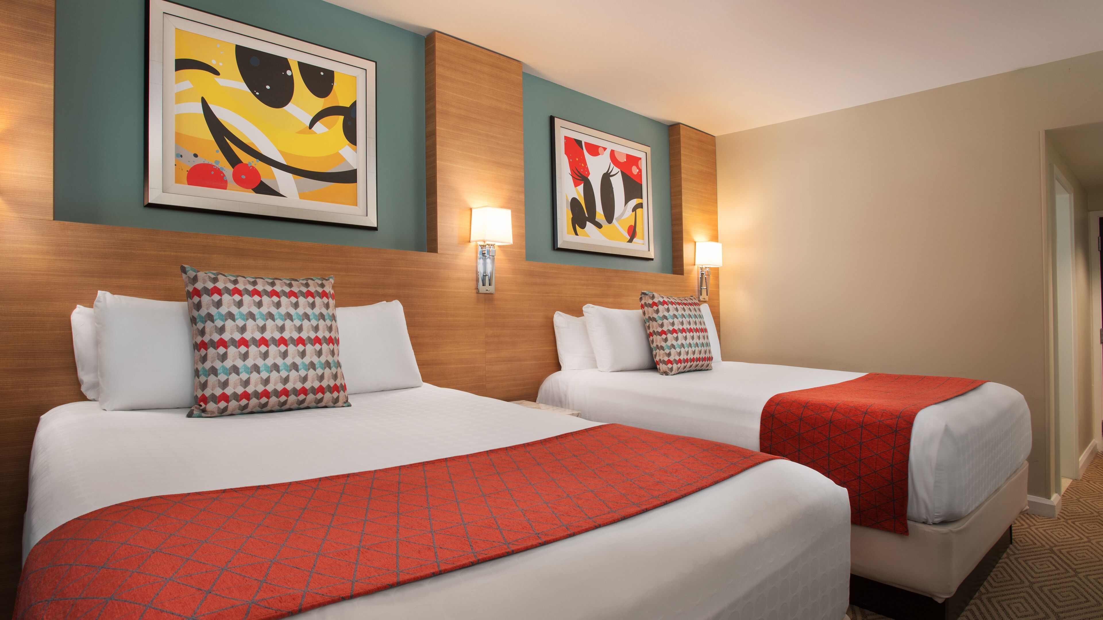 Quarto de hotel com duas camas, cada uma com almofada estampada e manta vermelha. Há quadros abstratos nas paredes e luminárias fixadas.