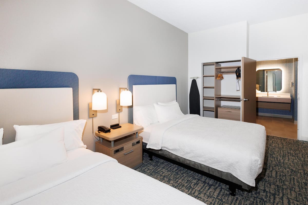 Habitación de hotel con dos camas, una mesita de noche con lámpara, un armario y un baño visible a través de una puerta abierta.
