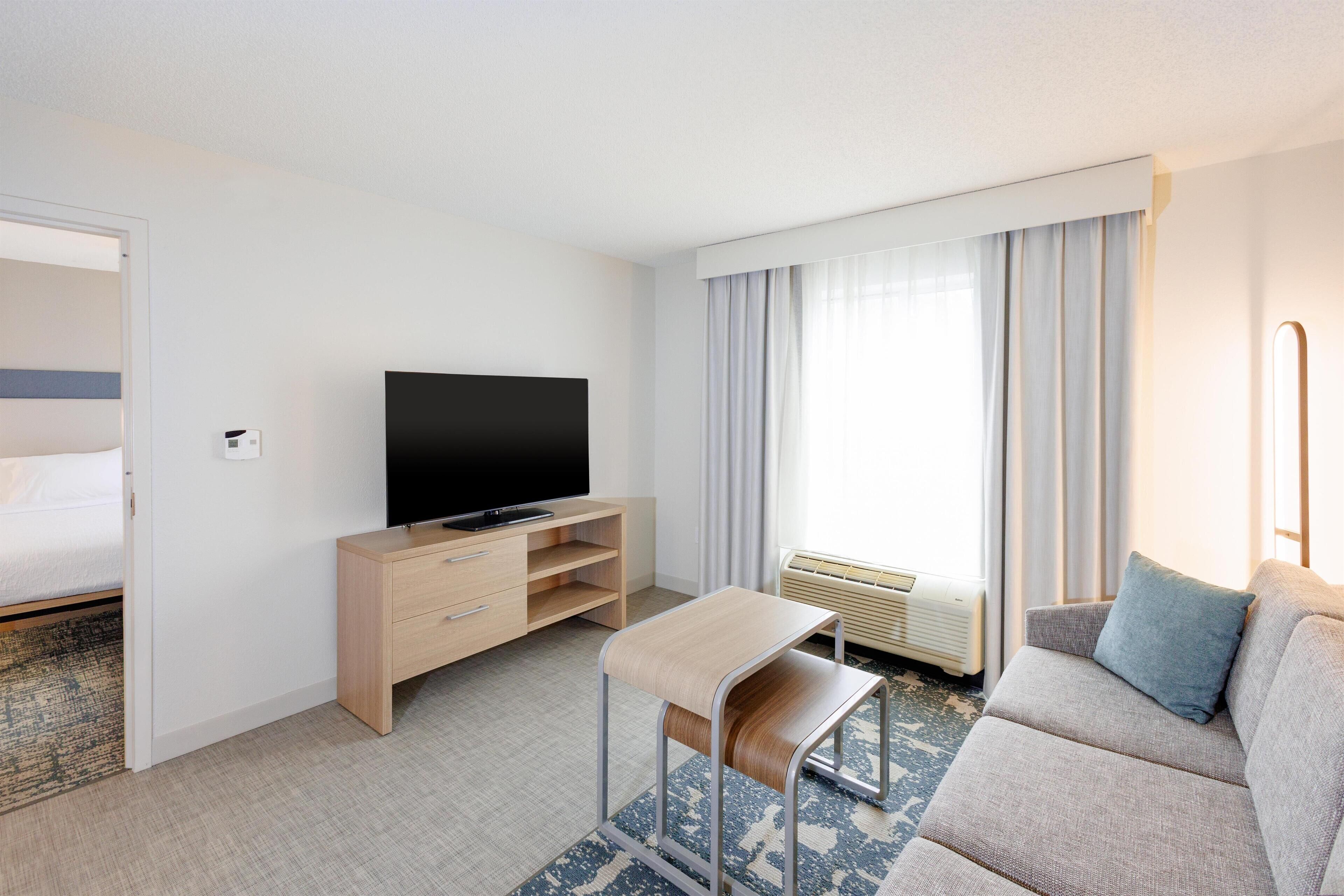 A hotel room with a bed, a TV, a sofa, a small table, and a window with curtains.