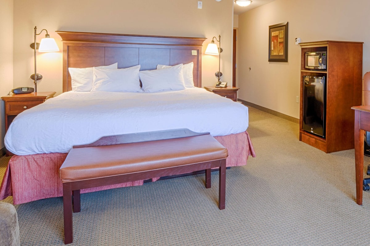 A large bed with white linens, a wooden headboard, two bedside lamps, a wooden nightstand, a wooden bench, a wooden wardrobe, and a microwave.