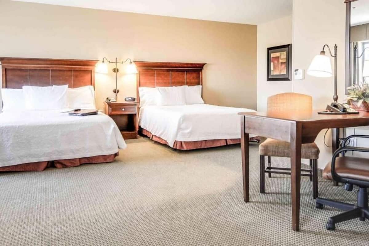 A hotel room with two beds, a desk, a chair, a lamp, and a mirror.