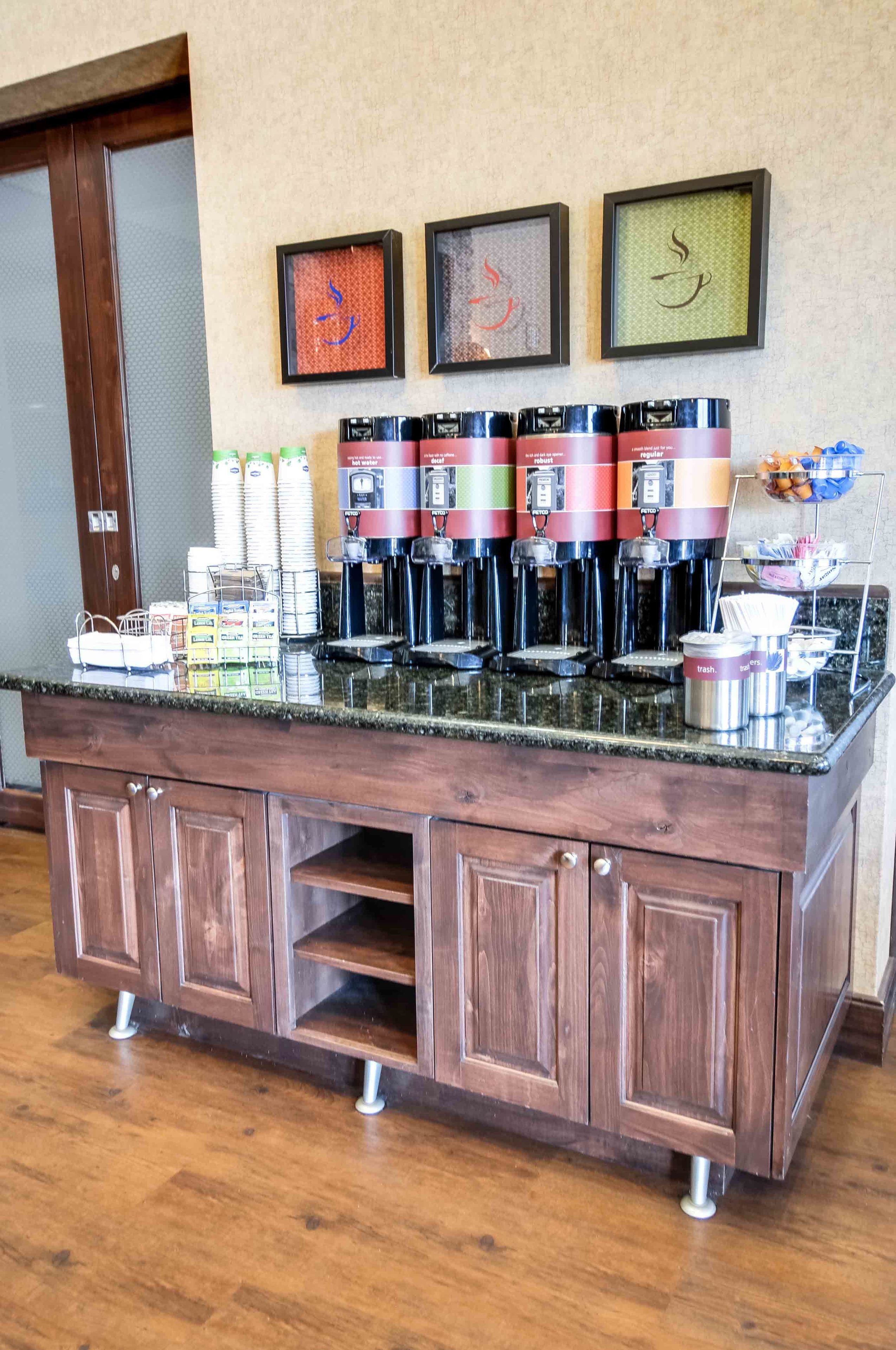 A coffee station with multiple coffee dispensers, a trash can, and a basket of snacks.