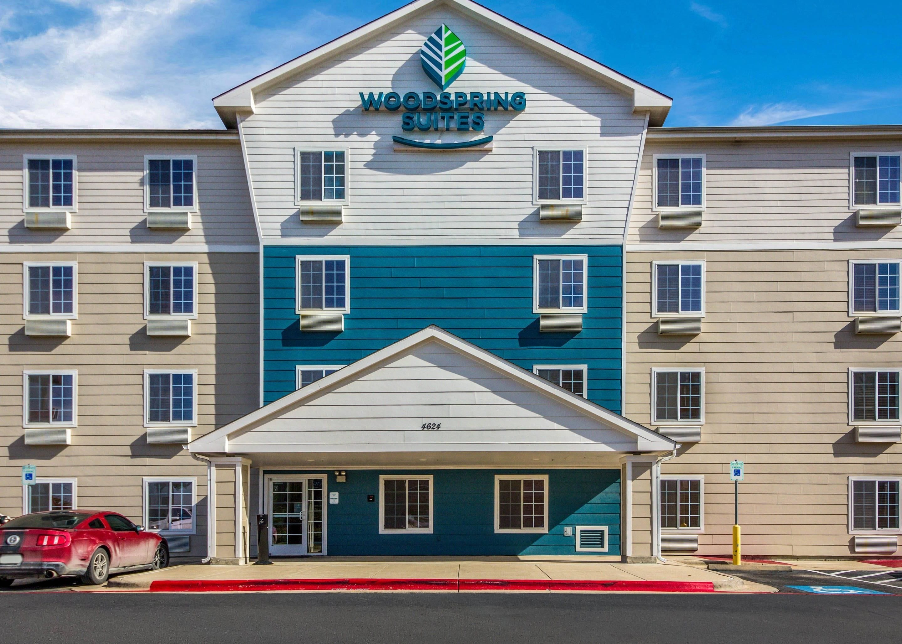 Um hotel com vários andares e um destaque para a placa 'Woodspring Suites', uma entrada coberta e uma faixa vermelha indicando uma área de estacionamento.