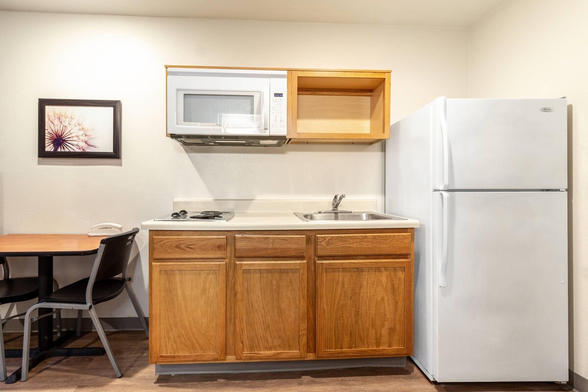 A compact kitchen with wooden cabinets, a white refrigerator, a microwave, a sink, and a chair.