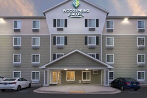 A multi-story hotel with a central entrance, air conditioning units, and a parking lot with cars.