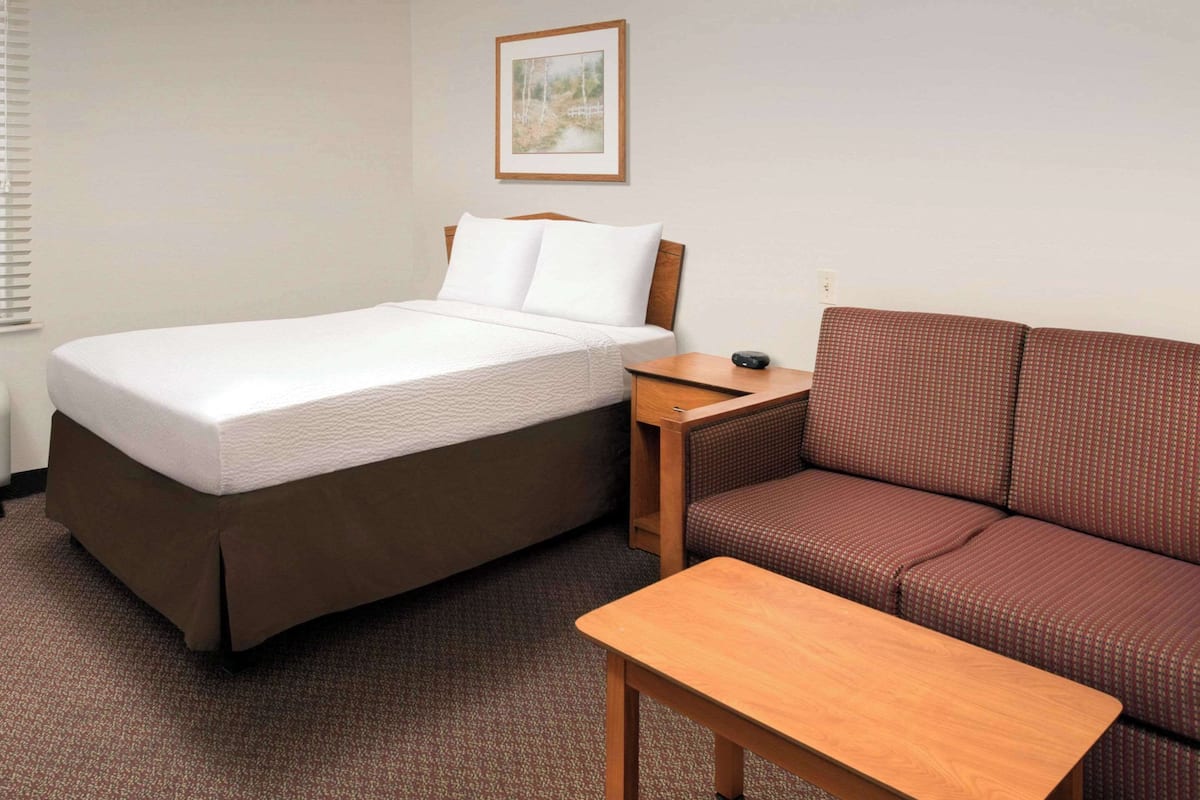 A hotel room with a bed, a wooden nightstand, a brown sofa, and a small wooden table.