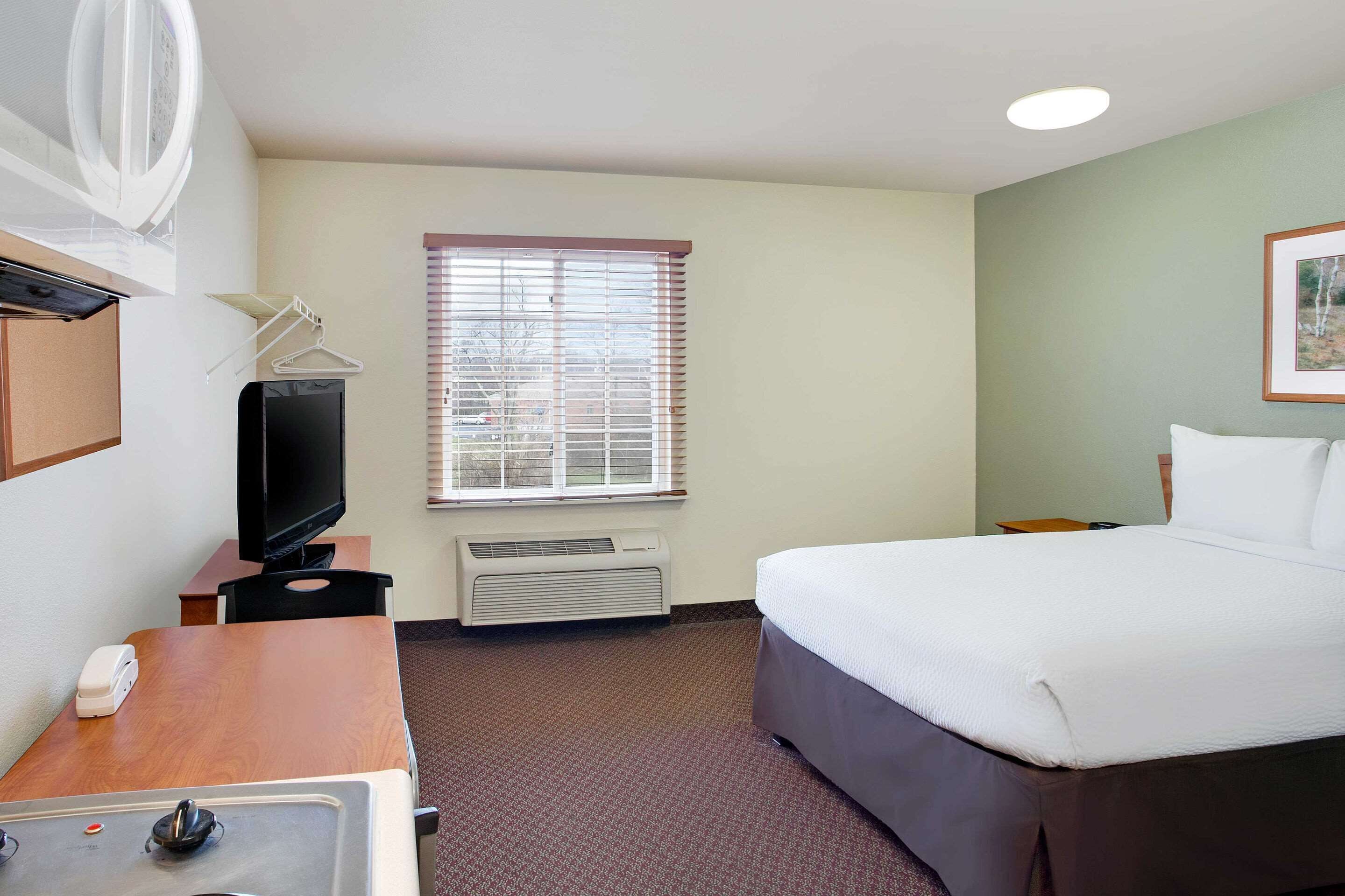 Habitación de hotel con cama, escritorio con teléfono, televisor, ventana con persianas y aire acondicionado instalado en la pared.
