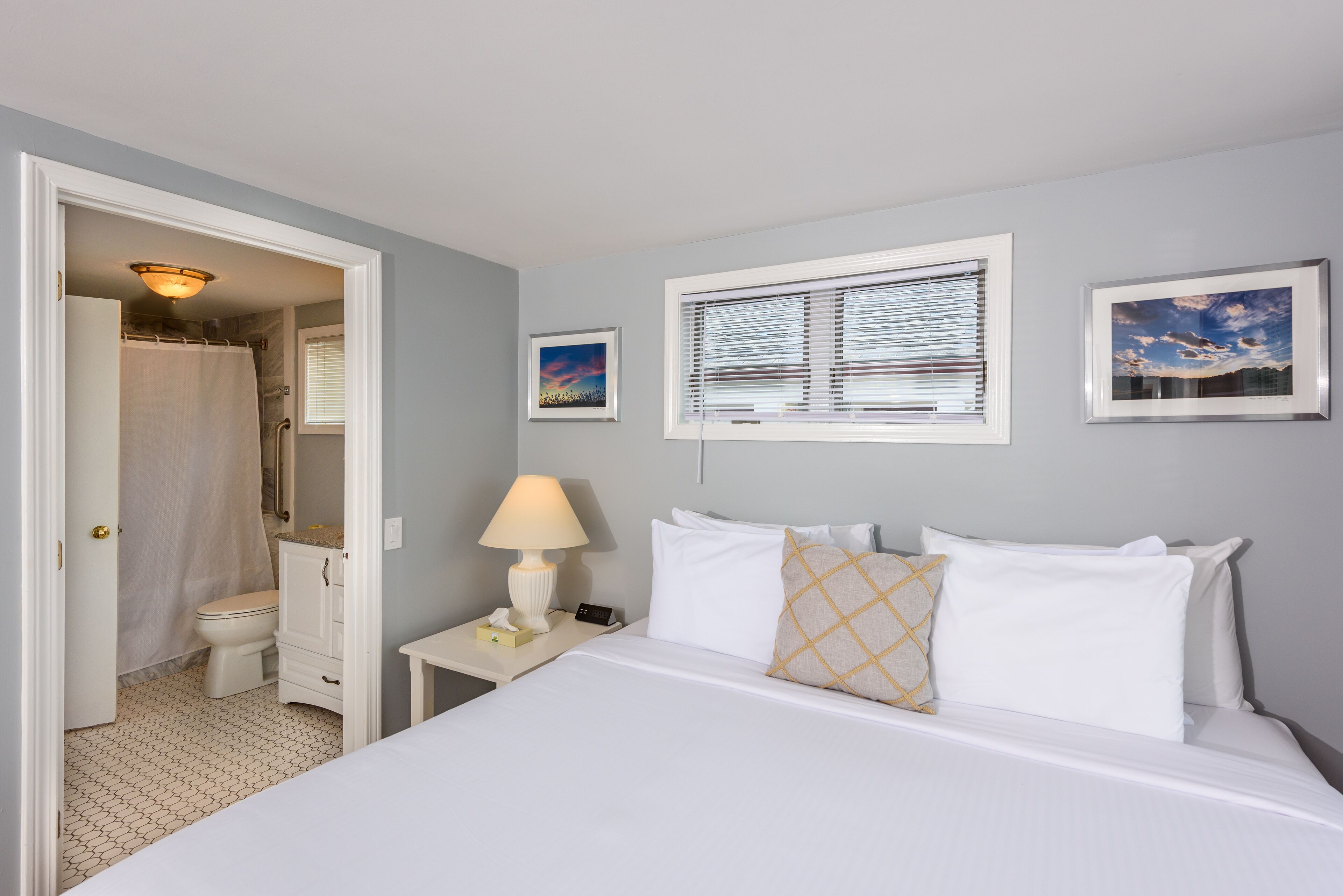 A bedroom with a bed, a nightstand with a lamp, a bedside table with a phone, a window with blinds, and a bathroom visible through an open door.
